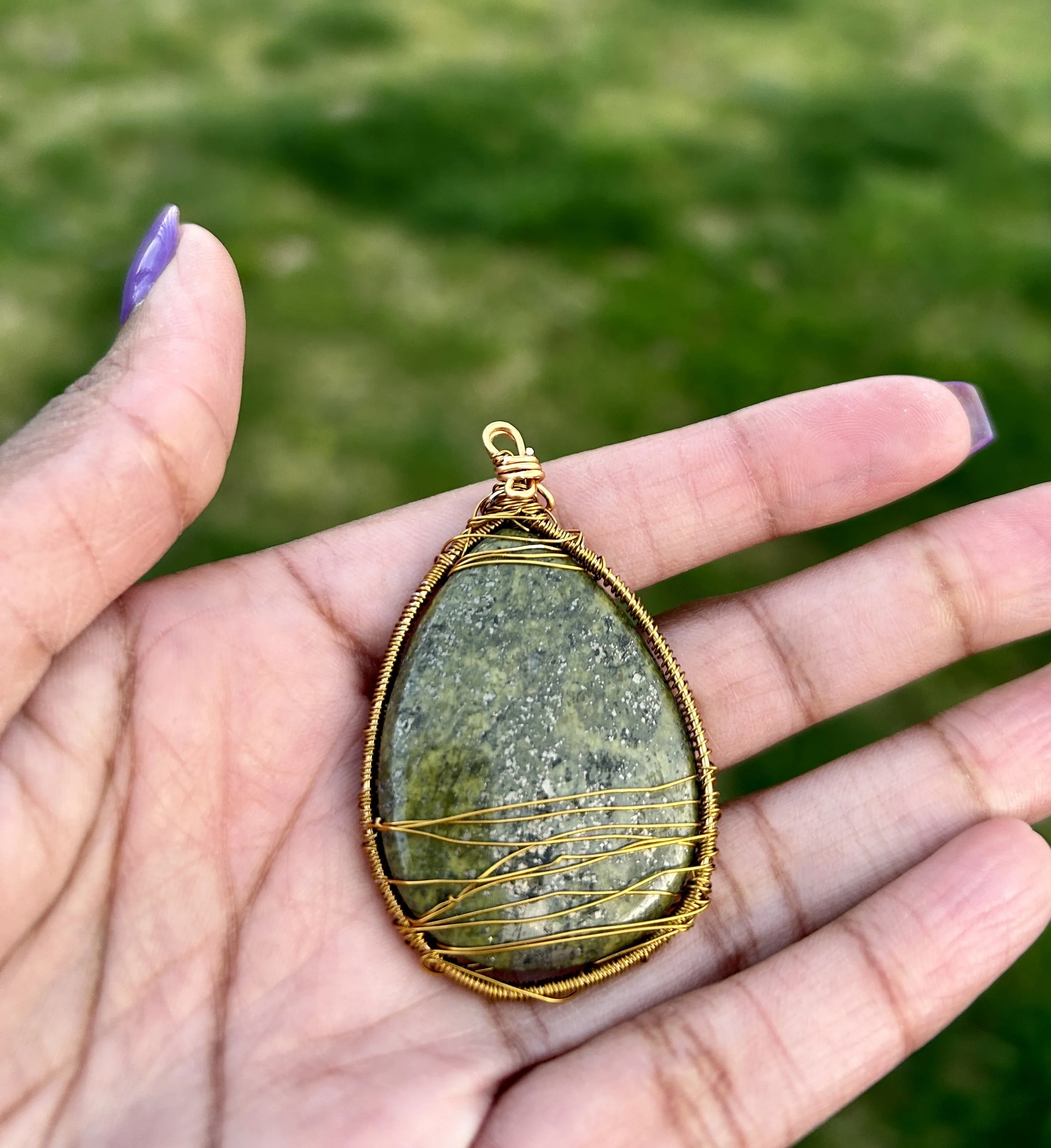 Hand holding a teardrop-shaped pendant with a green stone wrapped in gold wire, against a blurred green background.