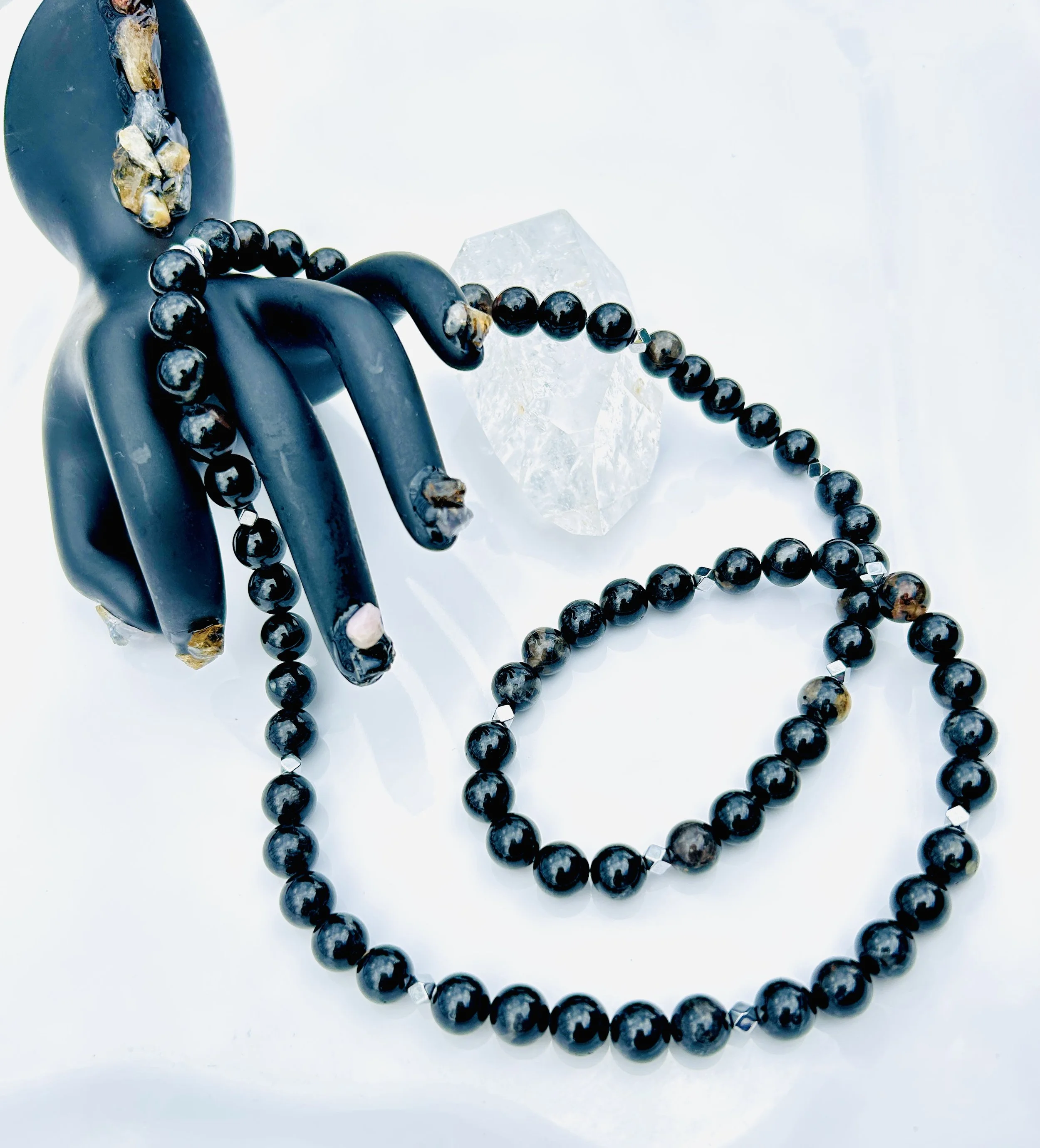 Black and gold beaded necklace draped over a black stone hand sculpture, with a clear quartz crystal in the background on a white surface.