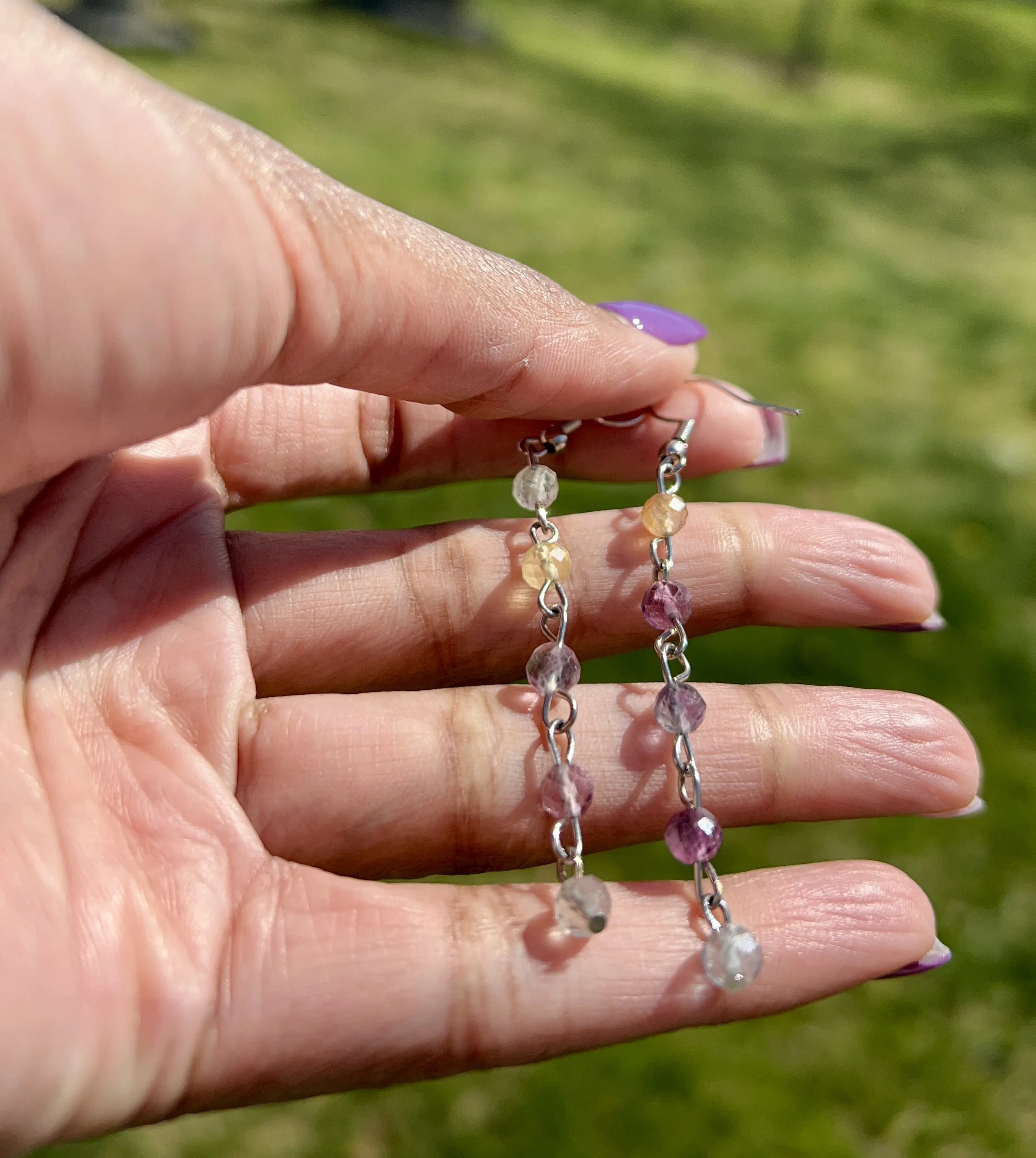 Close-up of a person's hand holding a pair of silver chain earrings adorned with multicolored small beads, with a green outdoor background.