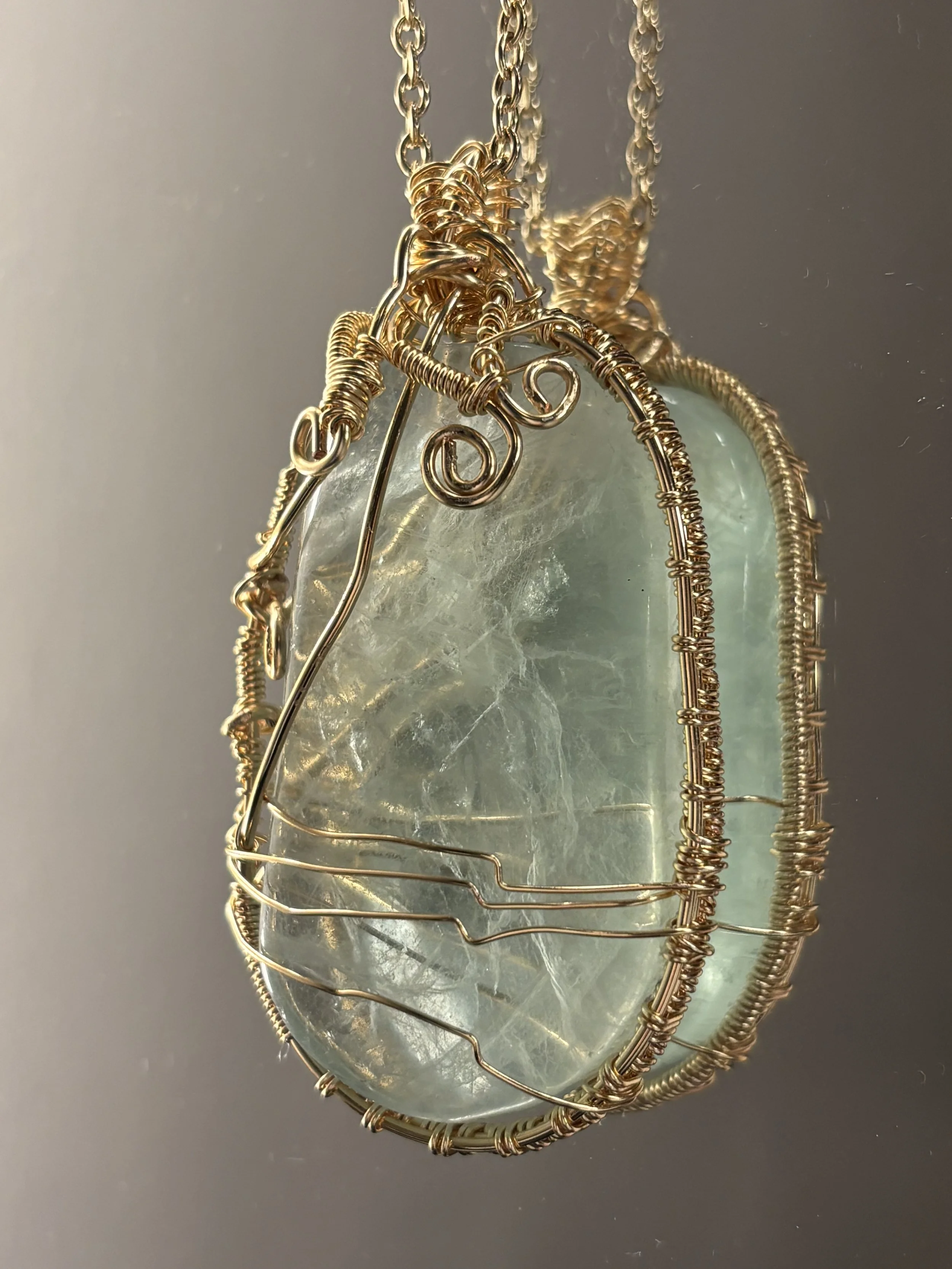A pendant with a large, irregularly shaped, greenish, semi-translucent gemstone wrapped in gold wire, hanging from a gold chain.