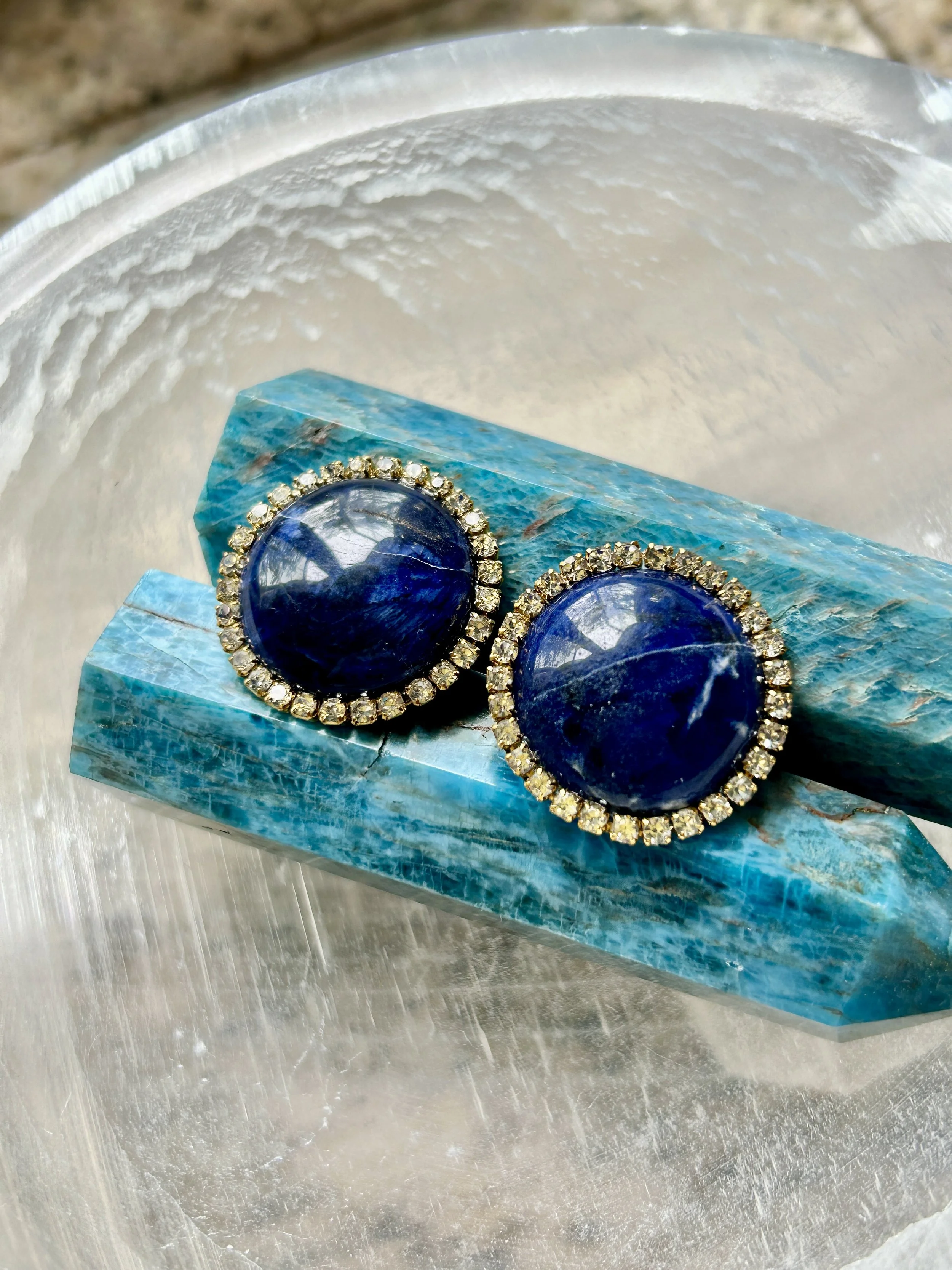 A pair of round navy blue earrings with a border of small clear stones resting on a textured blue stone slab.