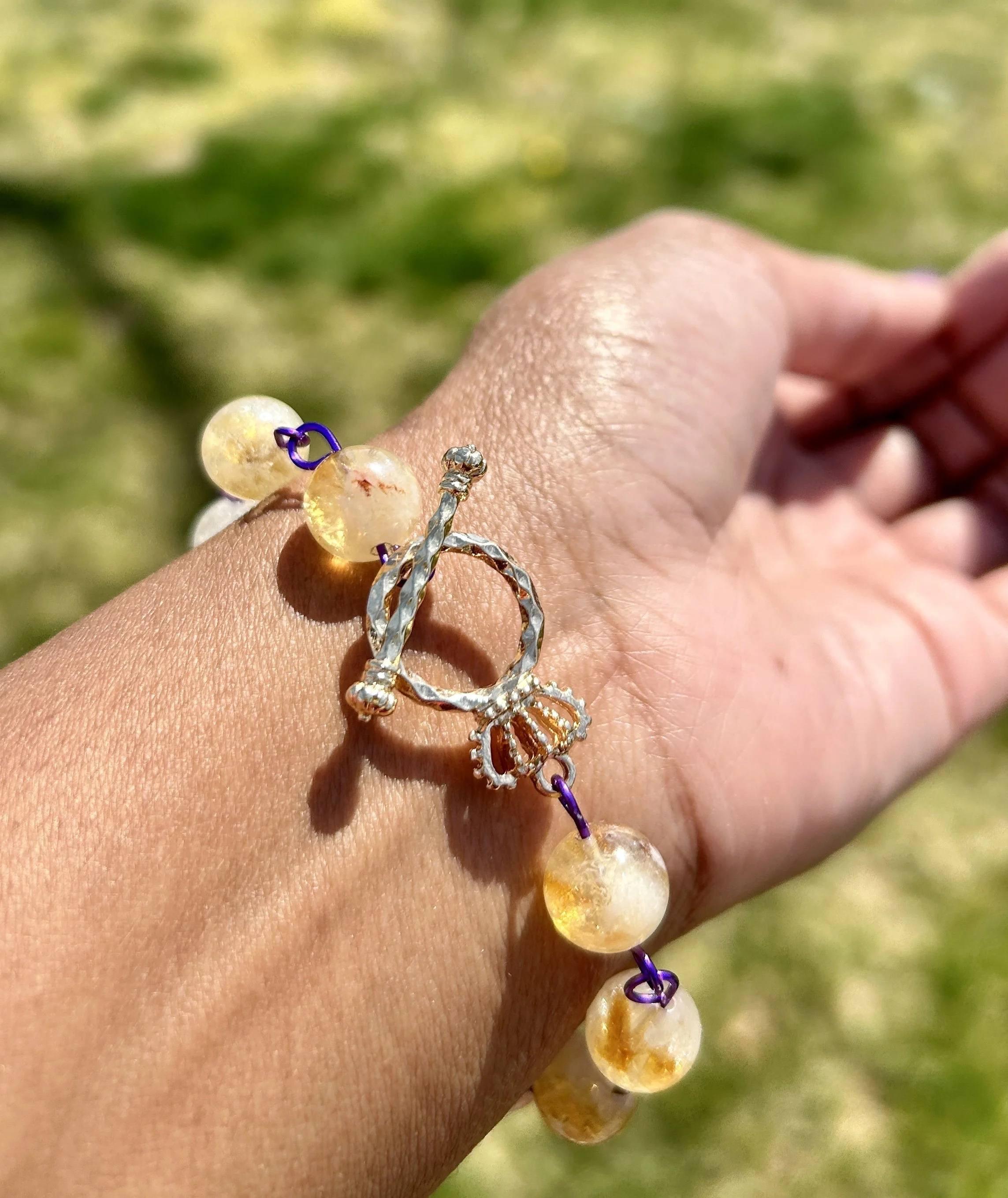 A bracelet with yellowish beads and a decorative charm held on a person's wrist, outdoors with a grassy background.