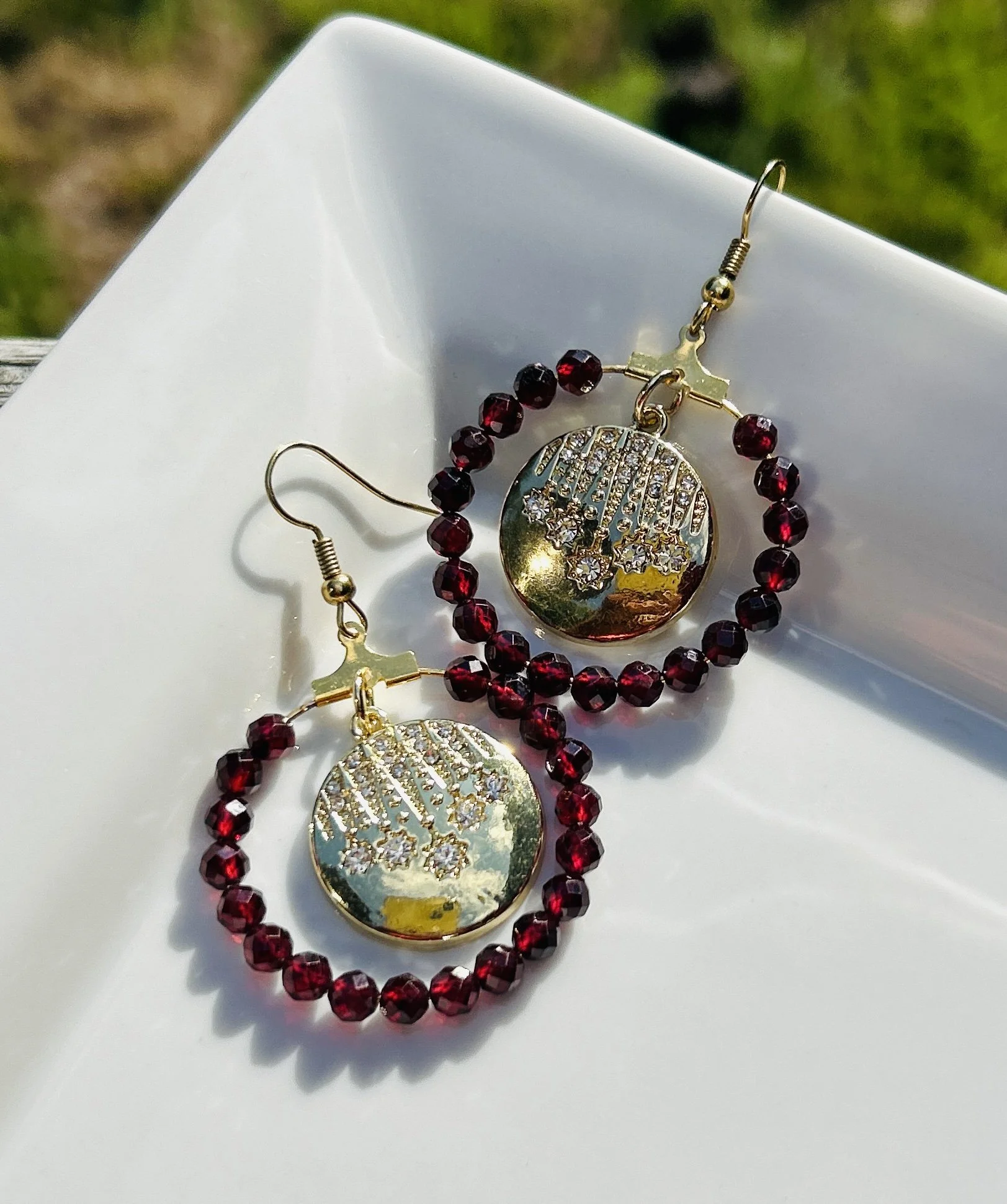 Pair of earrings with gold-colored discs inscribed with The Word, surrounded by dark red beads and placed on a white surface.