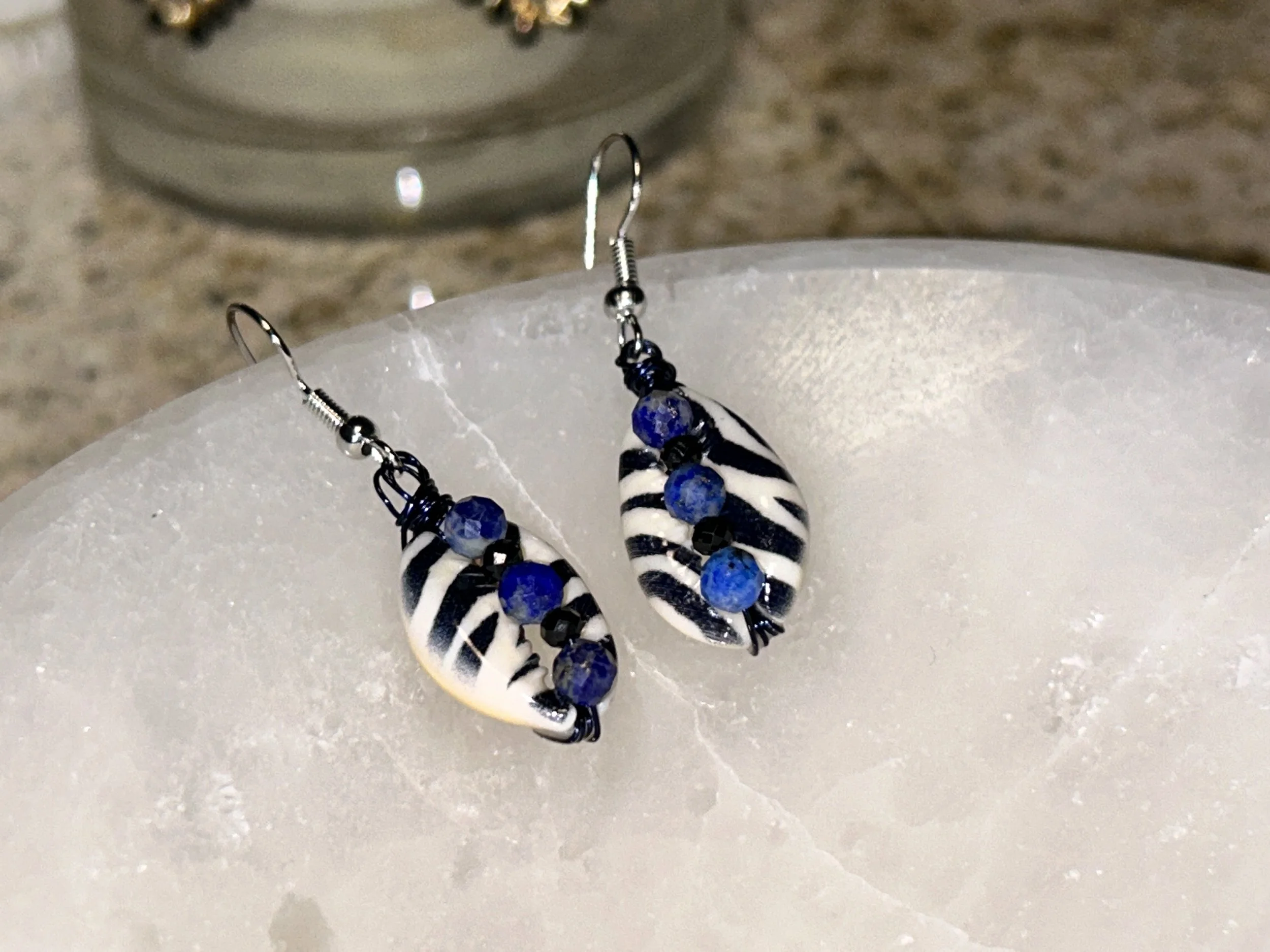 Pair of earrings with black and white striped beads and blue beads, hanging on white textured surface.