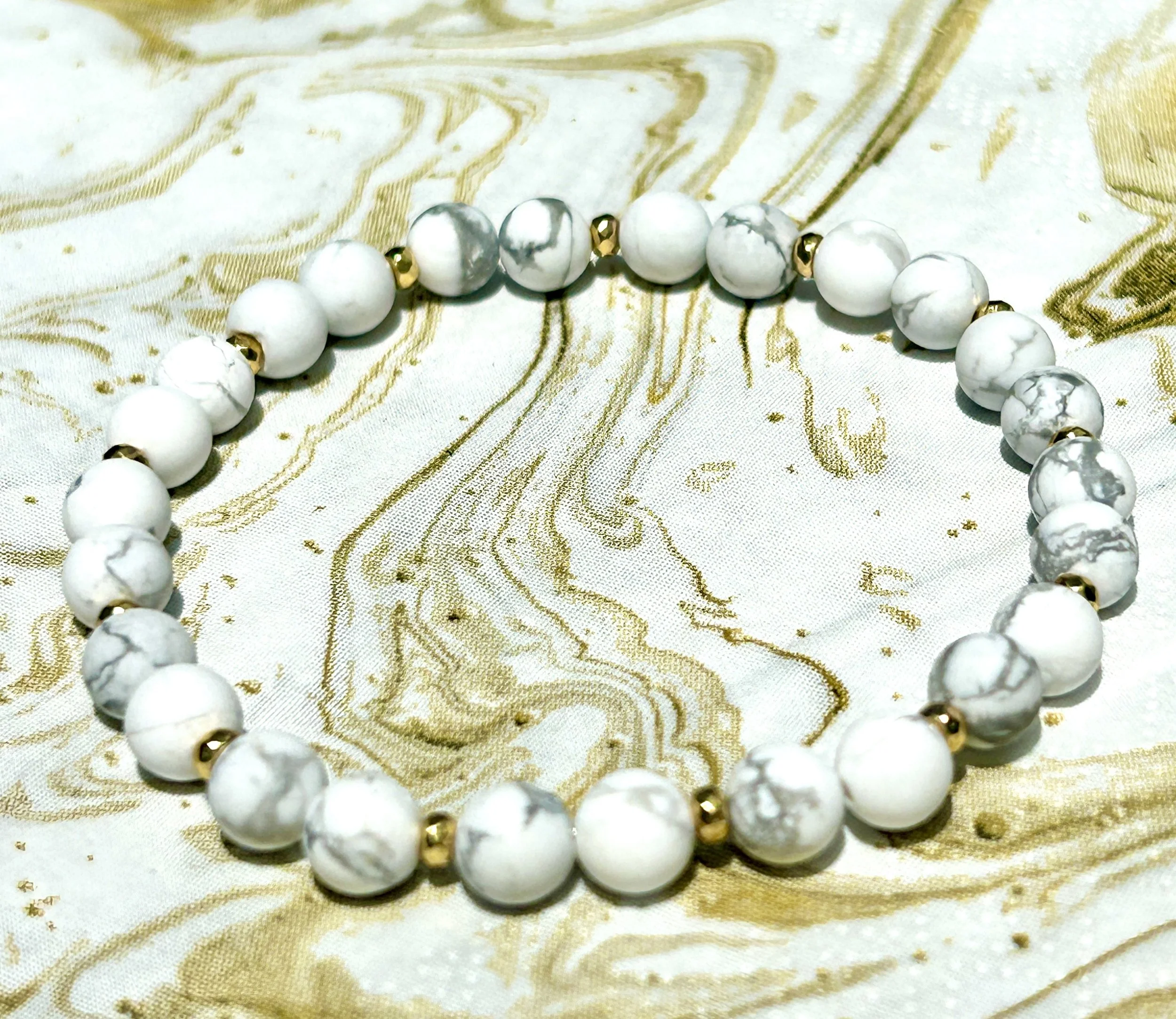 Beaded bracelet made of white marble-like round beads with small gold spacers, resting on a gold and white patterned fabric.