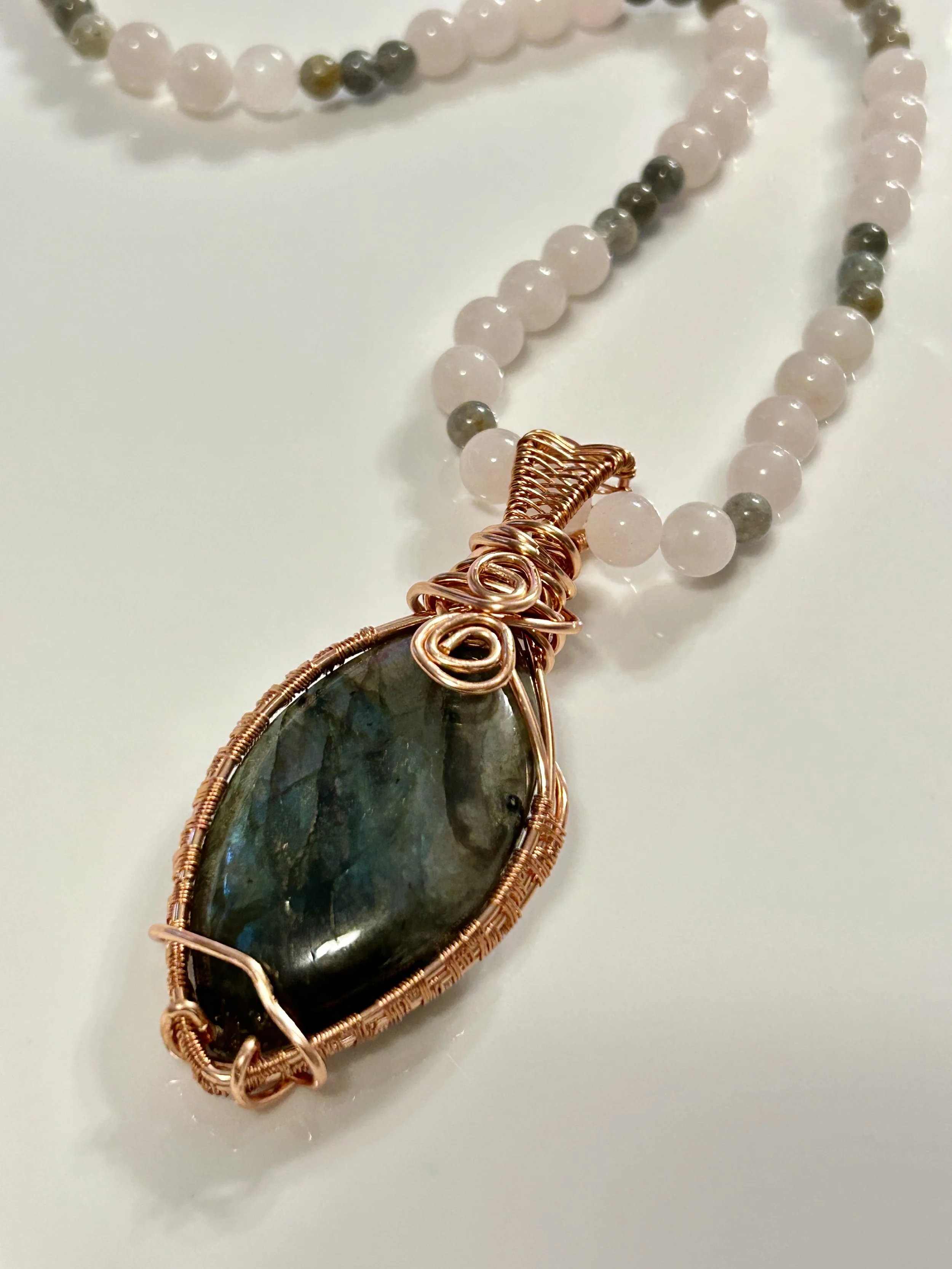 A beaded necklace with a large, dark, polished stone pendant wrapped in gold-colored wire, resting on a white surface.