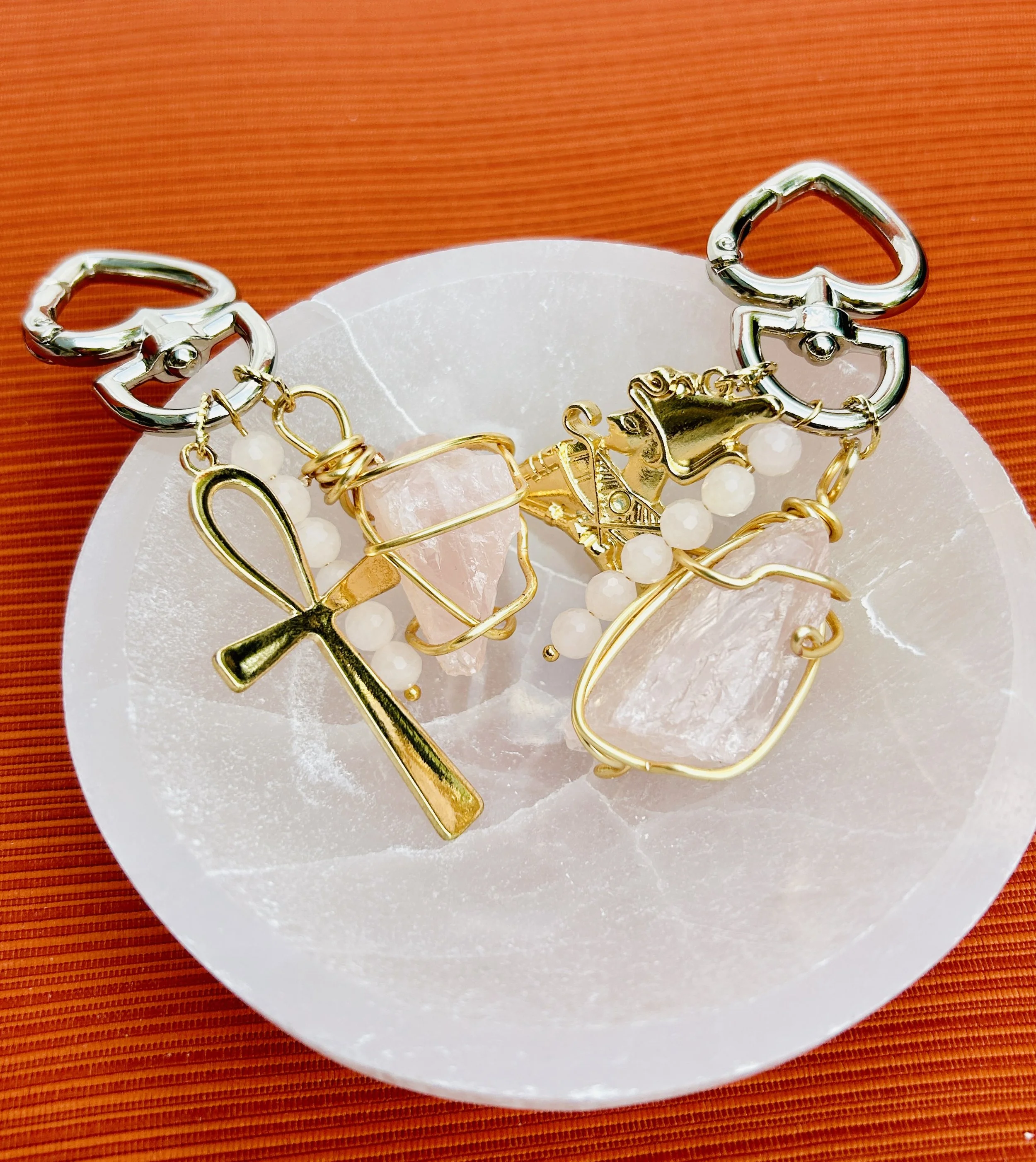Bracelet with various charms including an ankh, a unicorn head, and pink gemstone stones on white beads, placed on a white dish with an orange background.