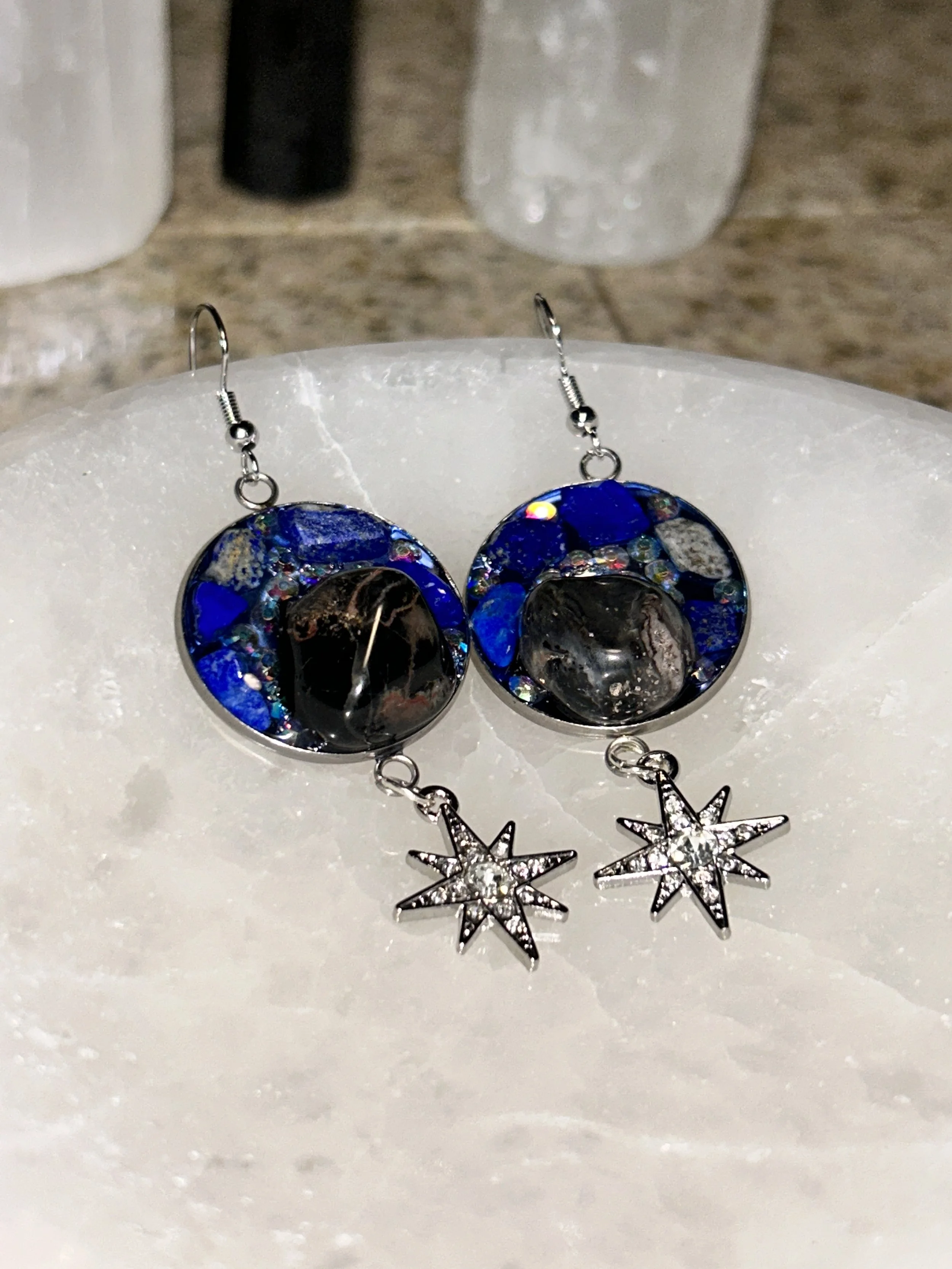 Pair of earrings with round black and blue gemstone inlays and star-shaped charms with small clear stones, displayed on a white surface.