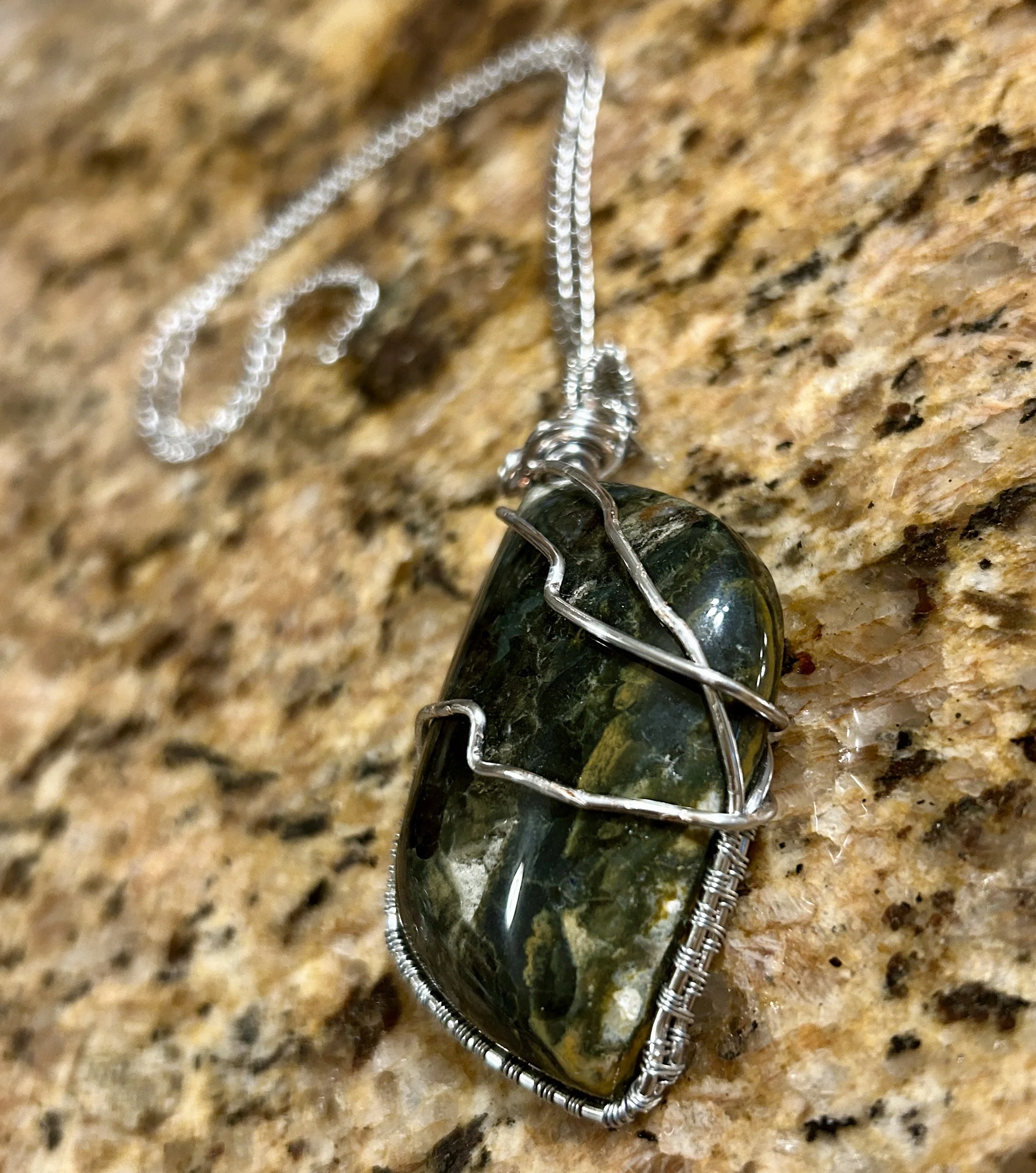 A pendant necklace with a large greenish stone wrapped in silver wire, resting on a granite countertop.