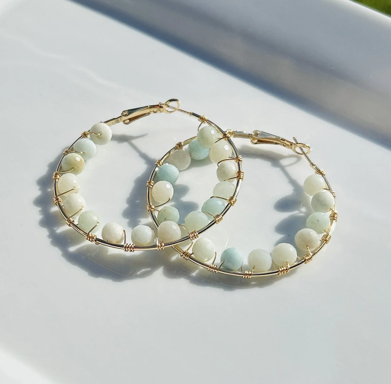 Gold hoop earrings with white and light blue beads, placed on a white surface with shadows cast by sunlight.