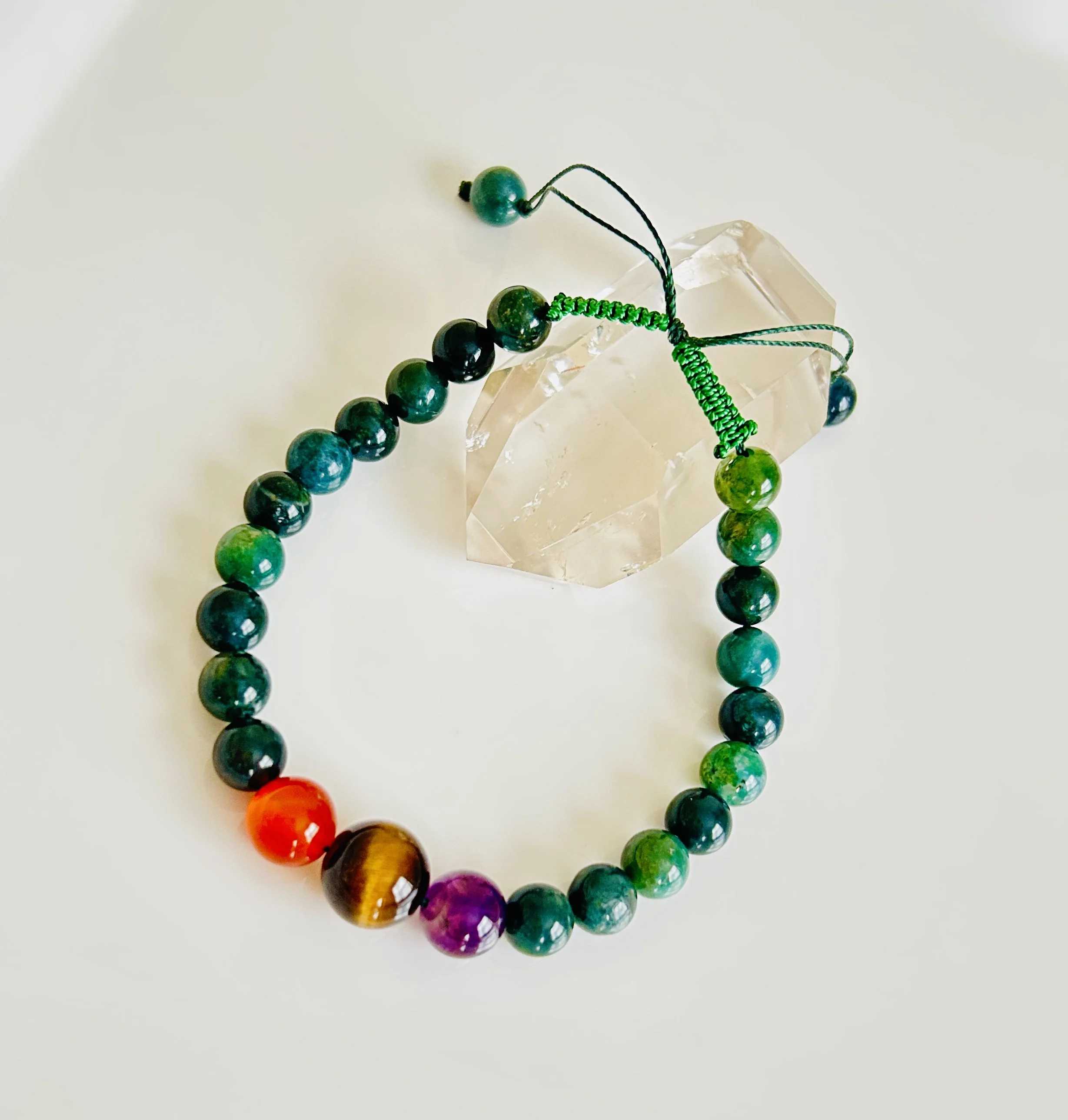 Colorful beaded bracelet with green, purple, orange, and tiger's eye beads, resting on a white surface next to a clear quartz crystal.