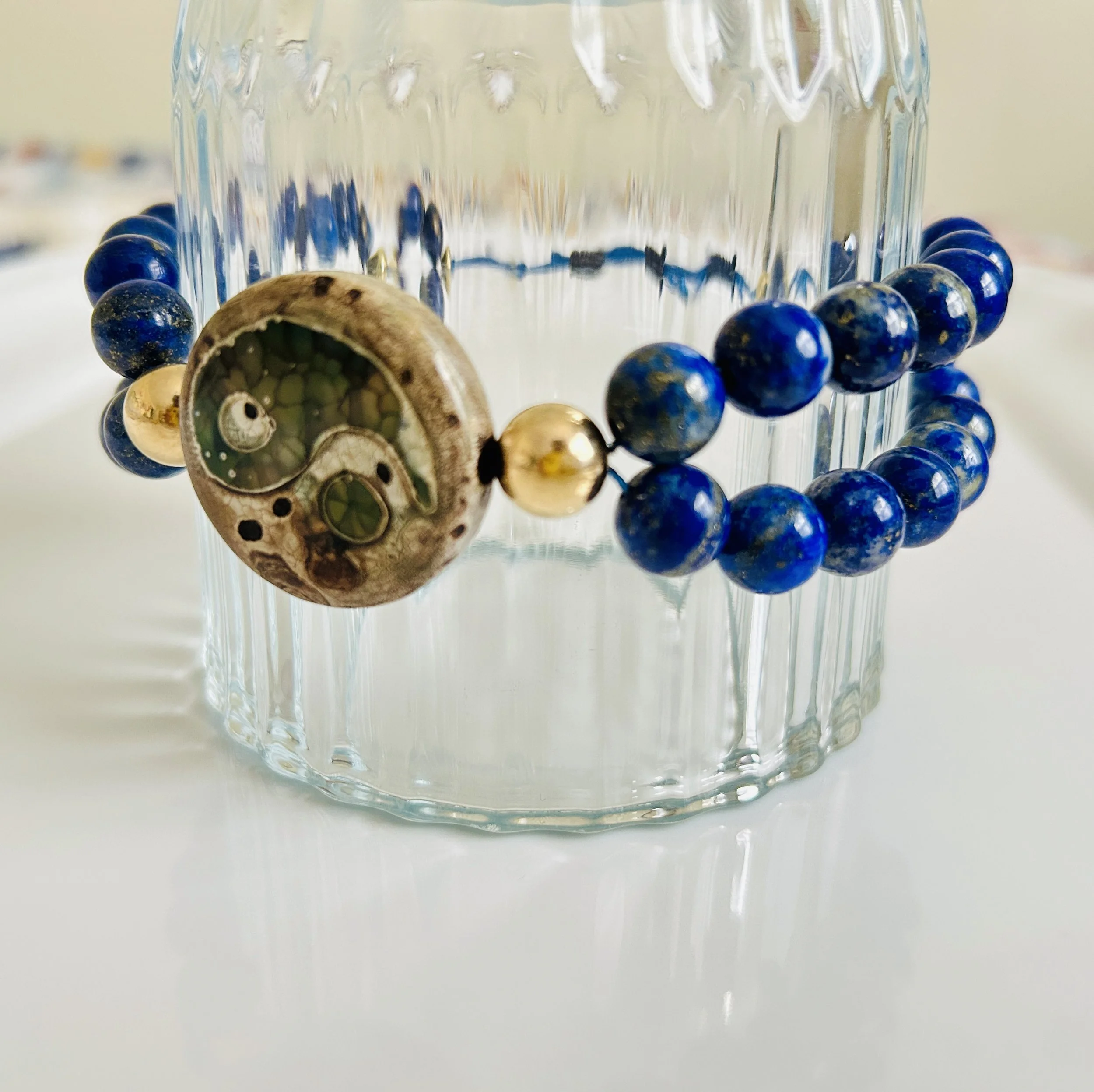A beaded bracelet with a large round pendant featuring a dial with gears and green accents, draped on a clear ribbed glass container.