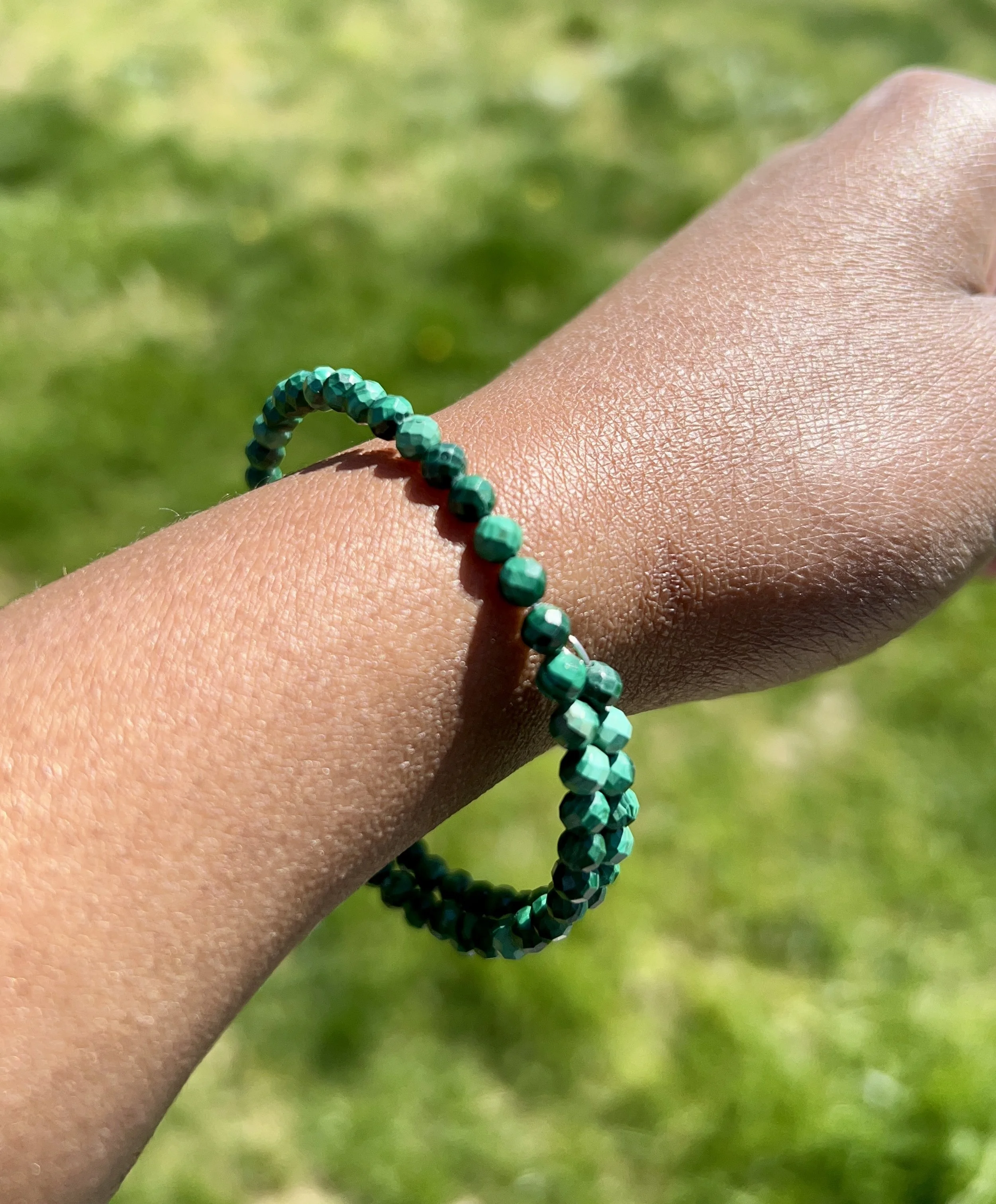A person's wrist wearing a green beaded bracelet with a grassy outdoor background.
