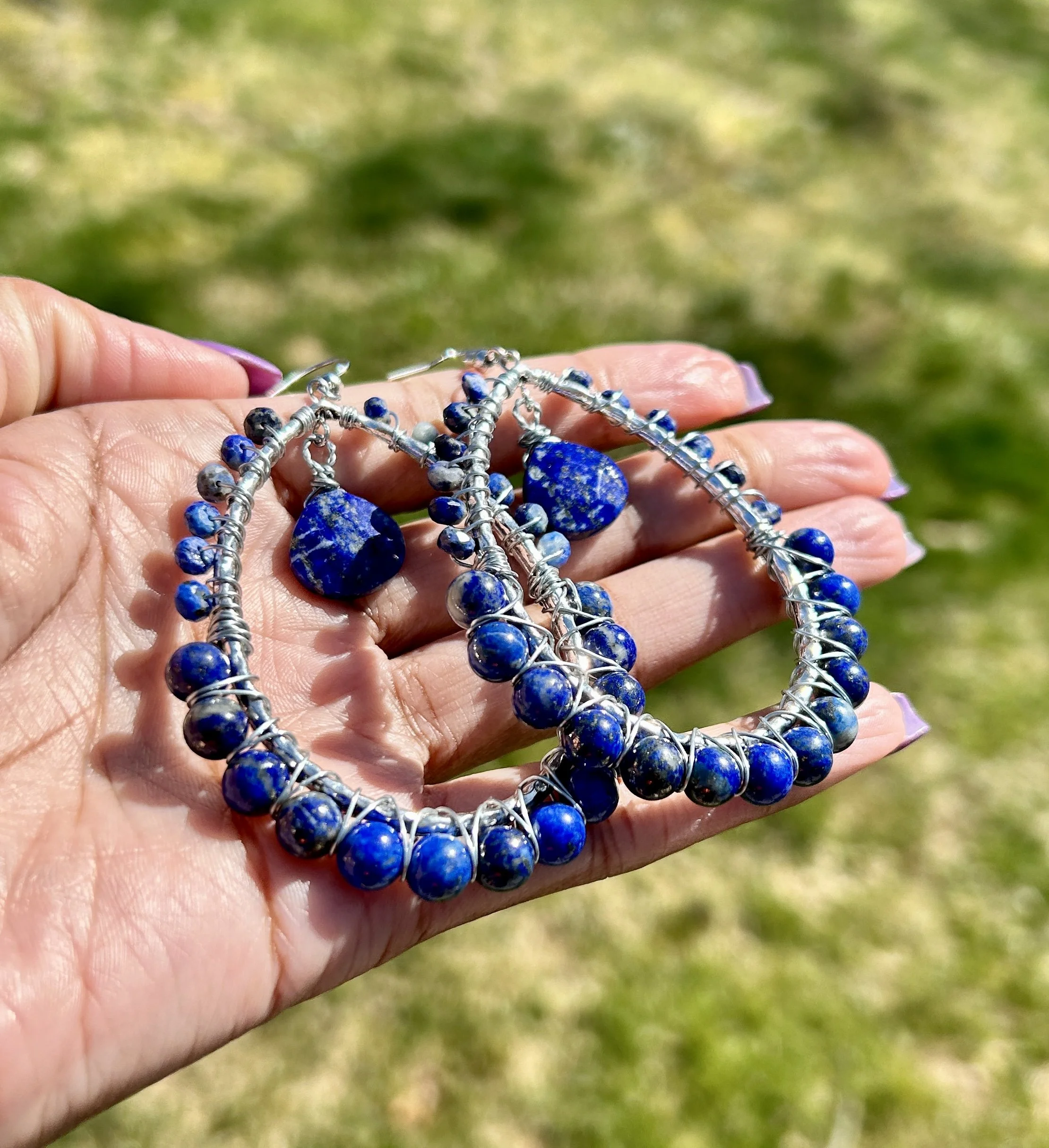 Hand holding a set of blue beaded jewelry, including earrings and bracelets, with a blurred outdoor grassy background.
