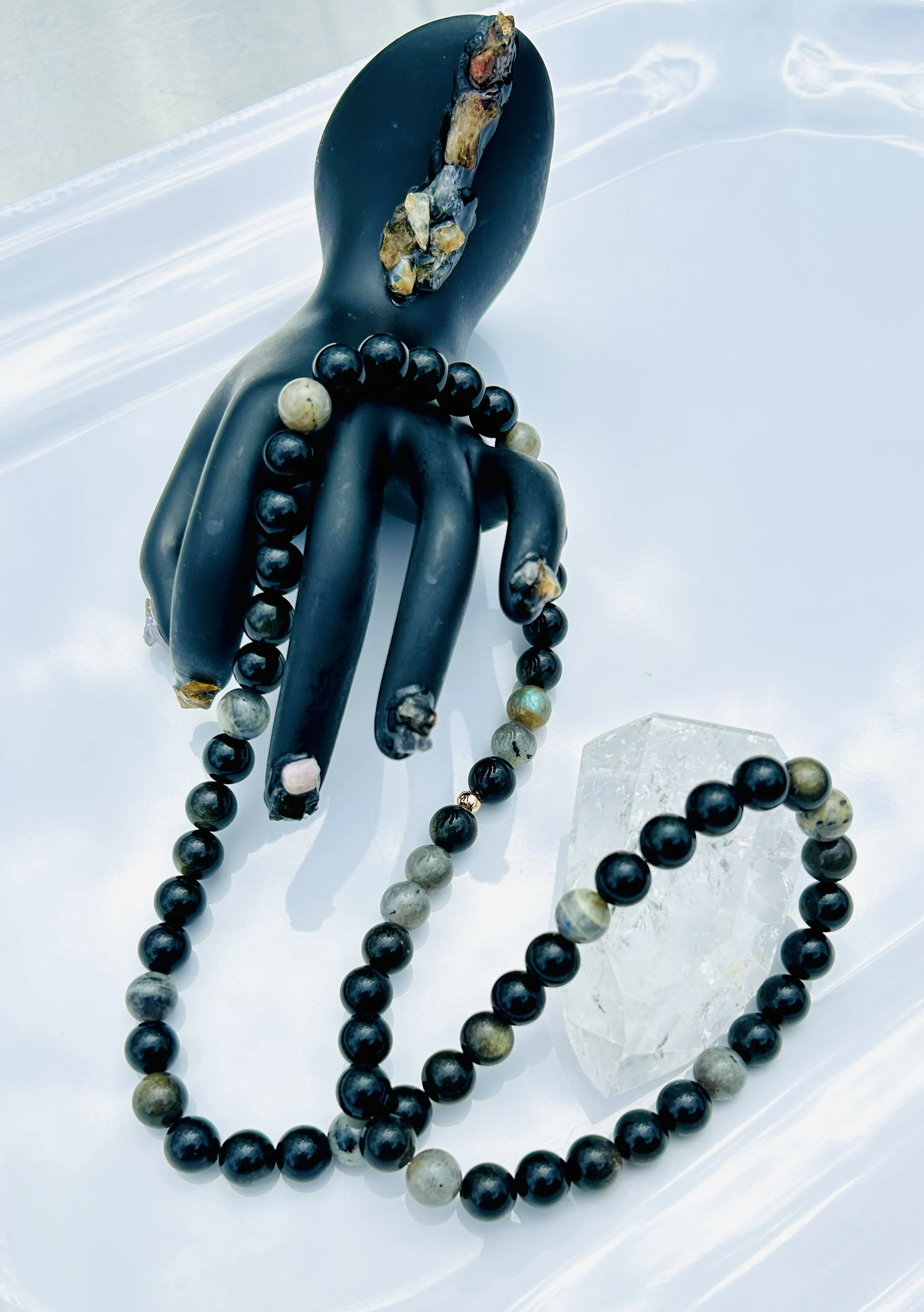 Decorative black hand sculpture with marble-like accents, draped with a beaded necklace made of black and gray beads, and a clear geometric resin piece on a white surface.