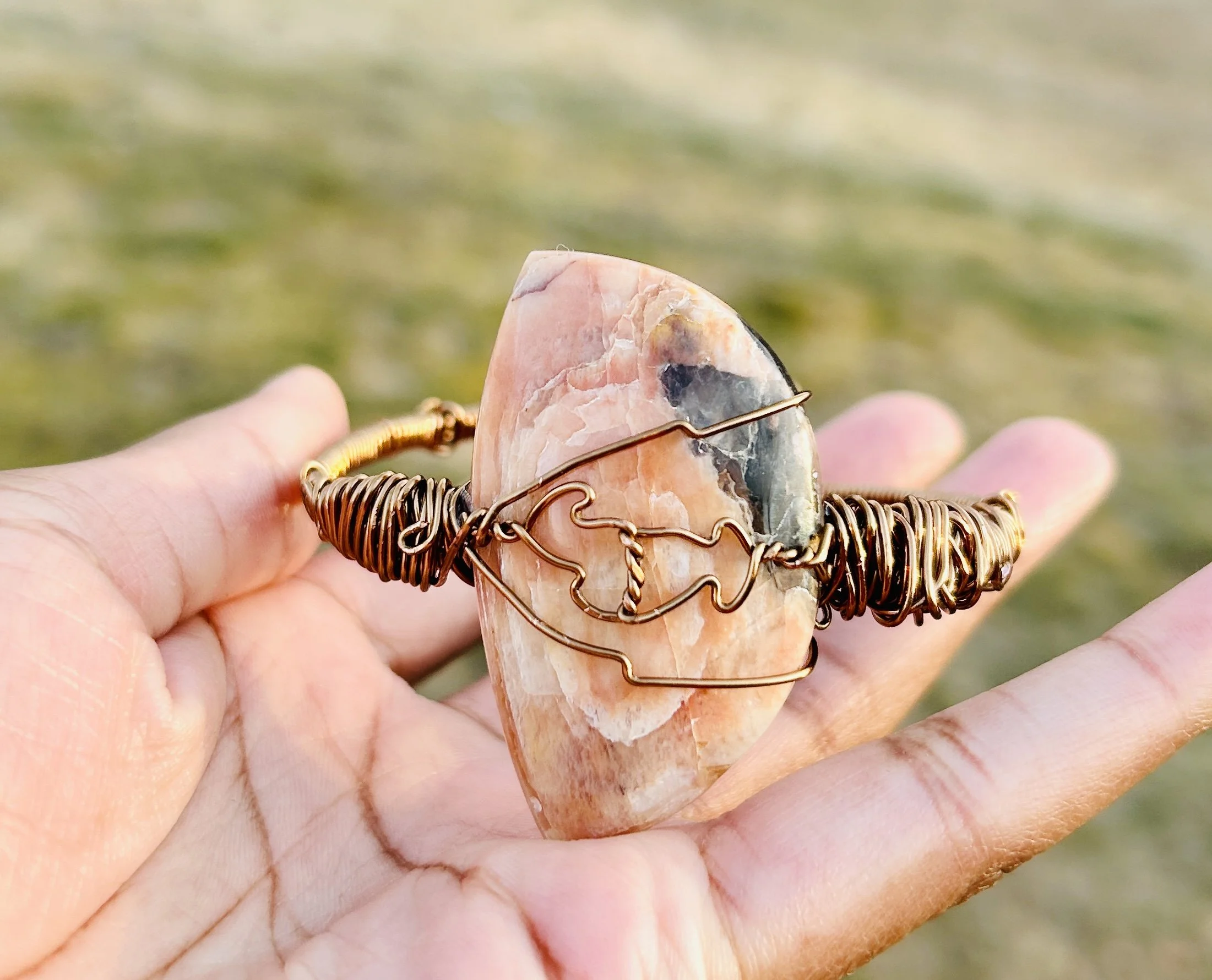 A hand holding a wire-wrapped bracelet with a large, flat, orange-pink stone against a blurred natural background.