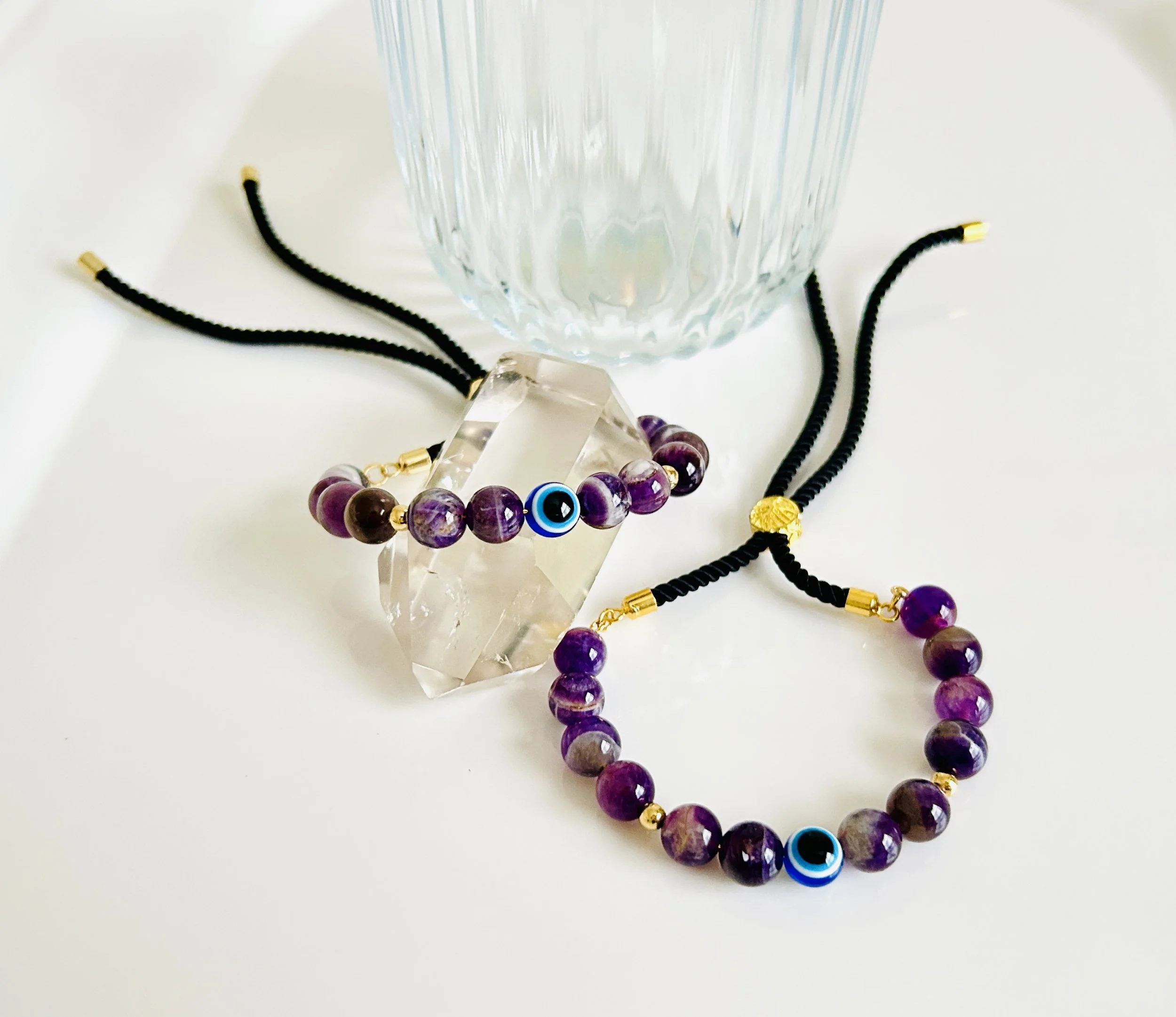 Jewelry set with purple beads, black cords, gold accents, and blue eye charms, arranged around a clear quartz crystal, with a glass bowl in the background on a white surface.