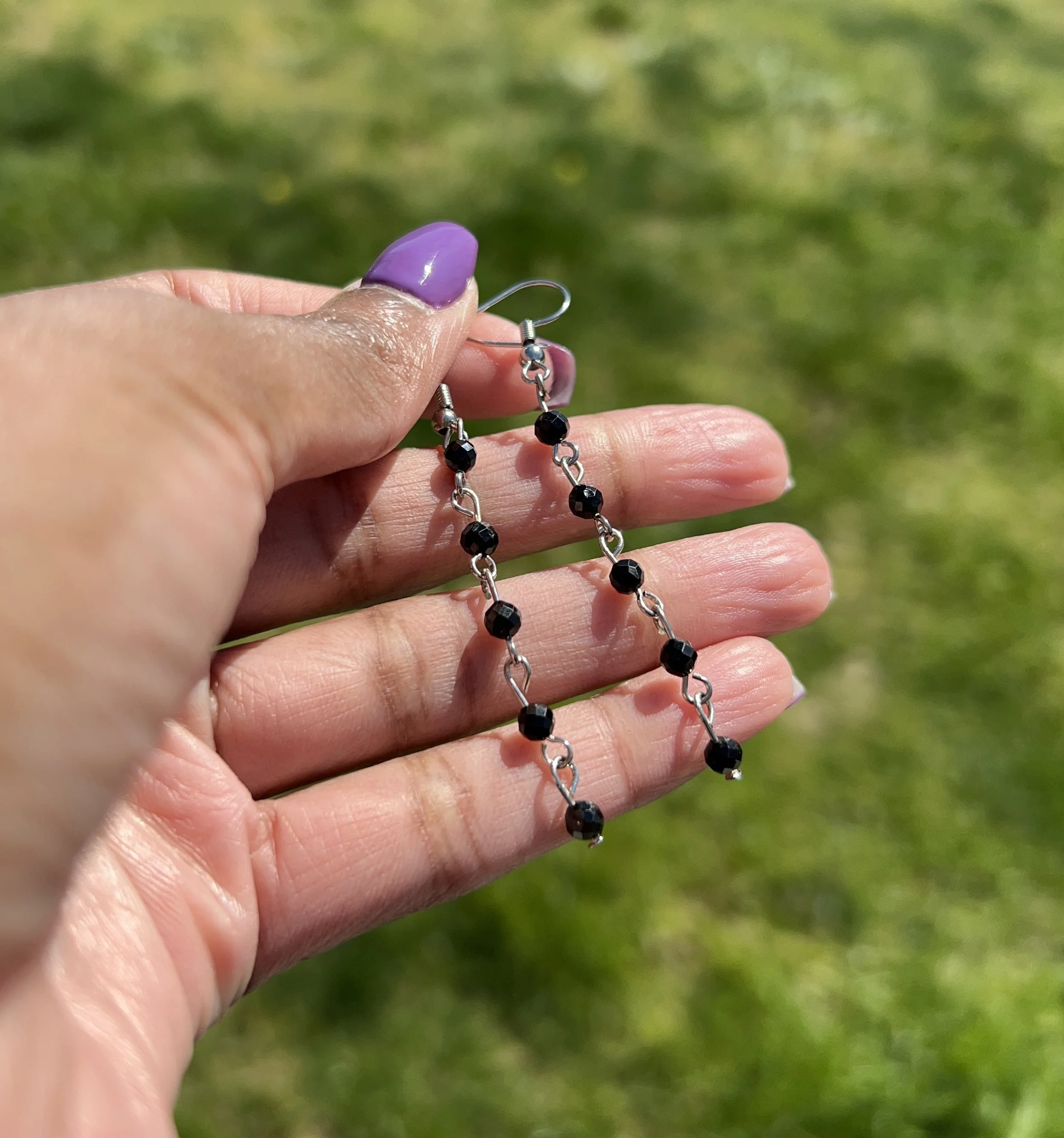 Hand holding black beaded chain earrings outdoors with grass background.