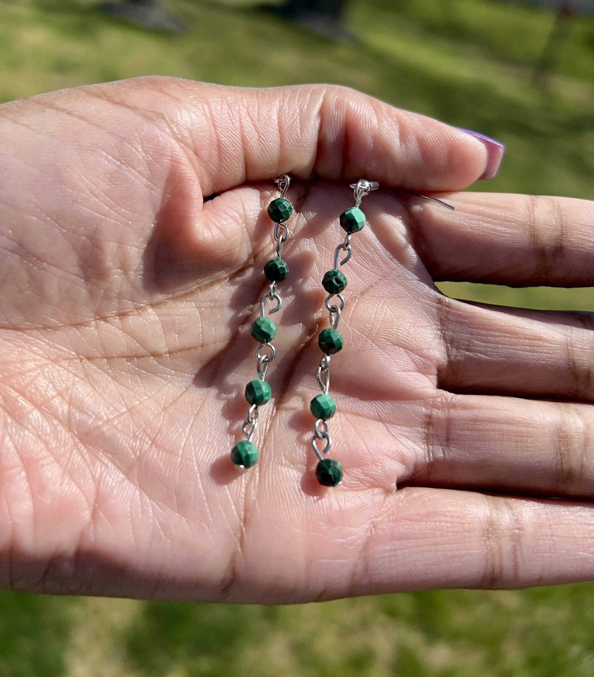 A person's hand holding a pair of long dangling earrings made of silver chains with dark green beads, with a grassy outdoor background.
