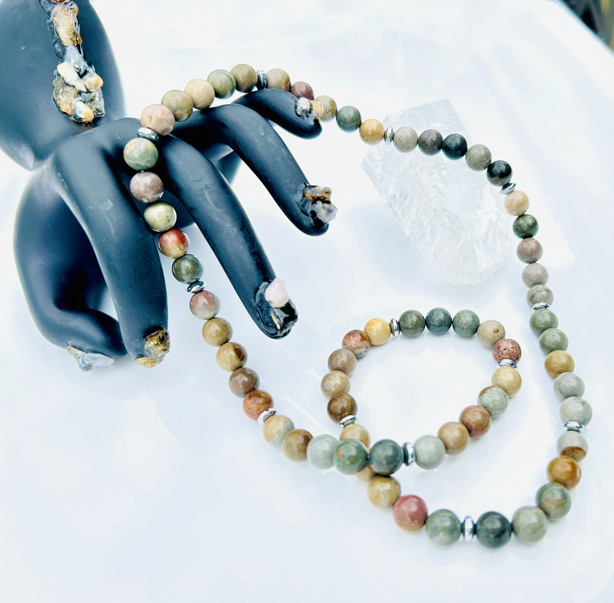 A multicolored beaded necklace and bracelet draped over a black ceramic hand sculpture, with a clear glass-like block and a white background.