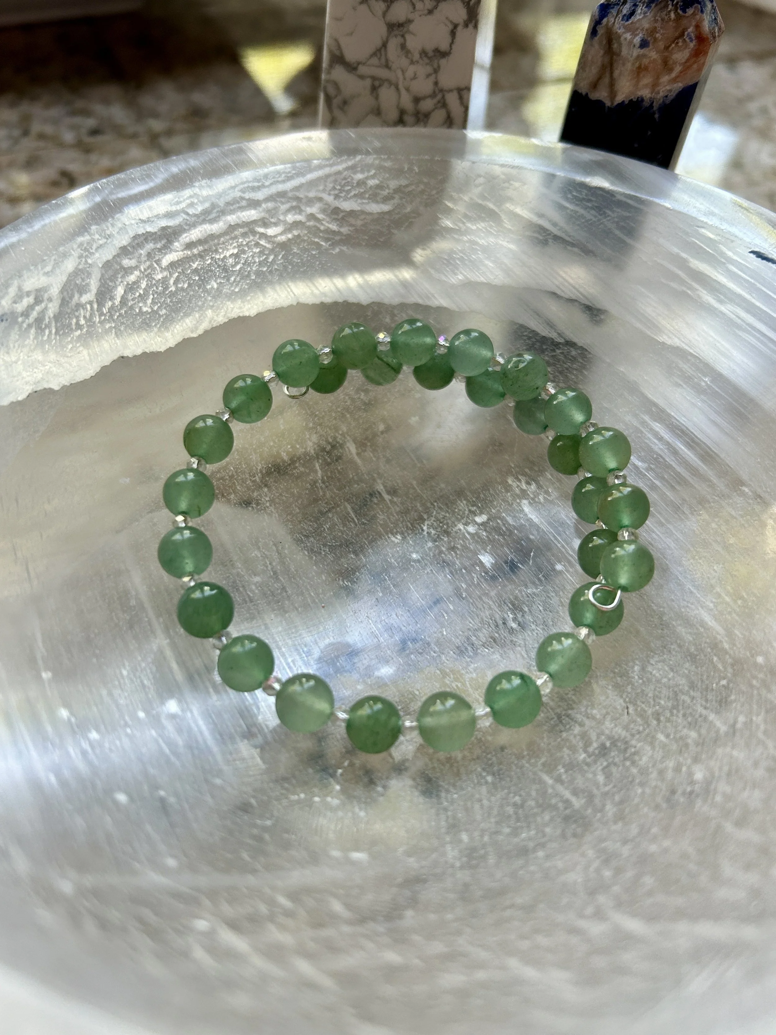 A green beaded bracelet resting inside a silver bowl. Two art prints are in the background.