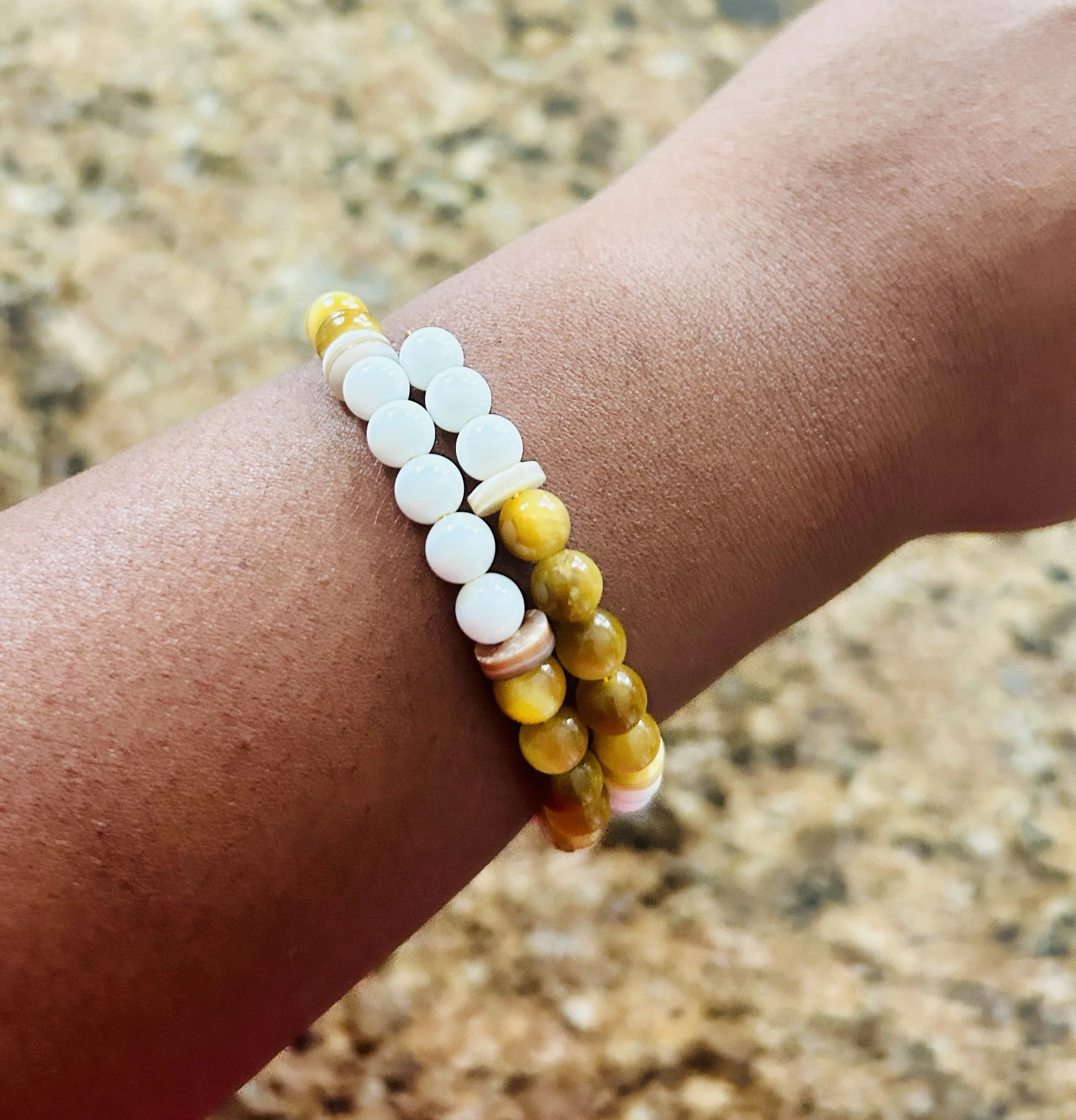 A person's wrist wearing two beaded bracelets, one with white beads and the other with yellow and brown beads, against a granite countertop background.