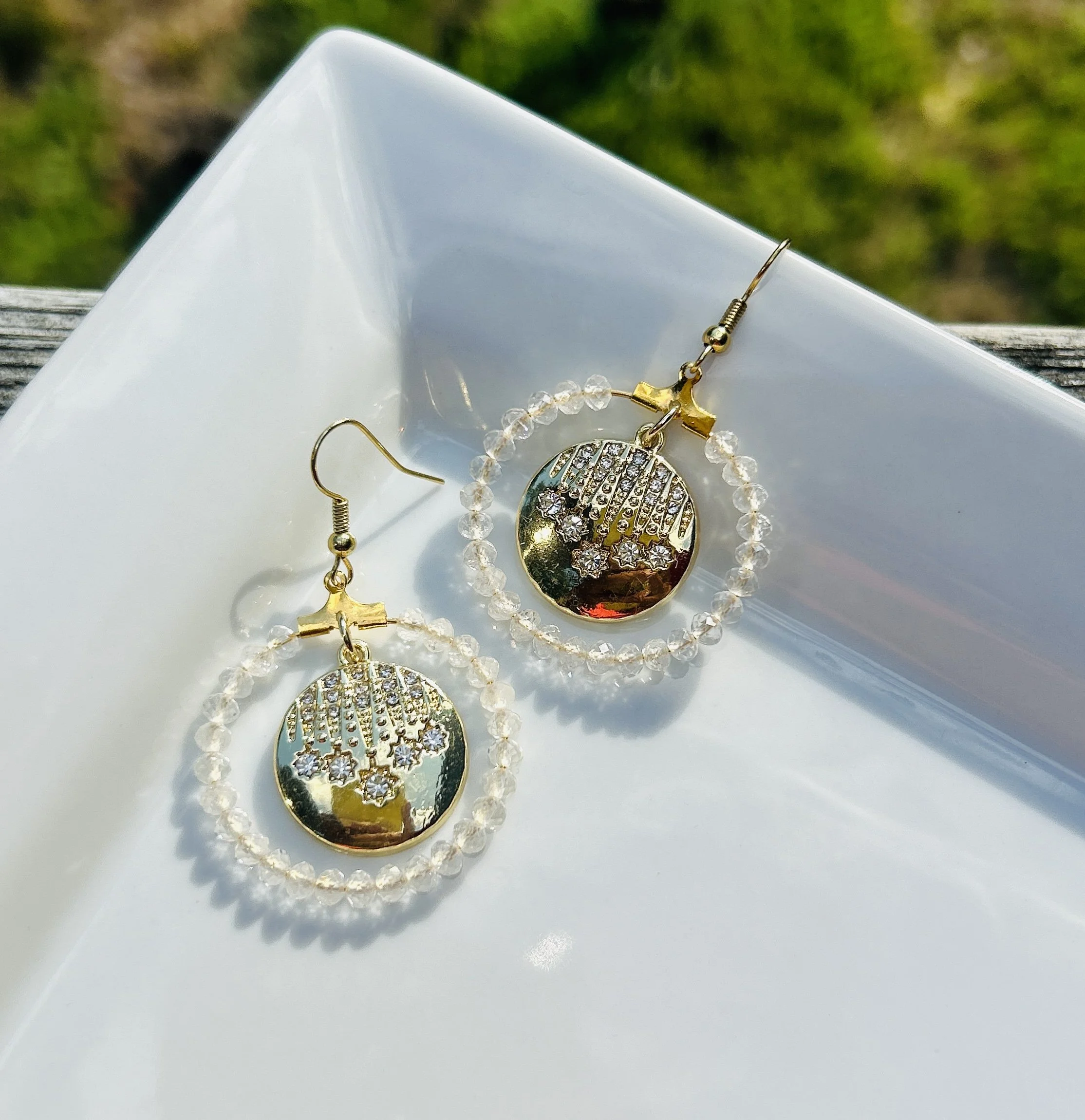 Gold dangling earrings with circular pendants surrounded by small clear beads, adorned with star-shaped rhinestones, placed on a white tray.