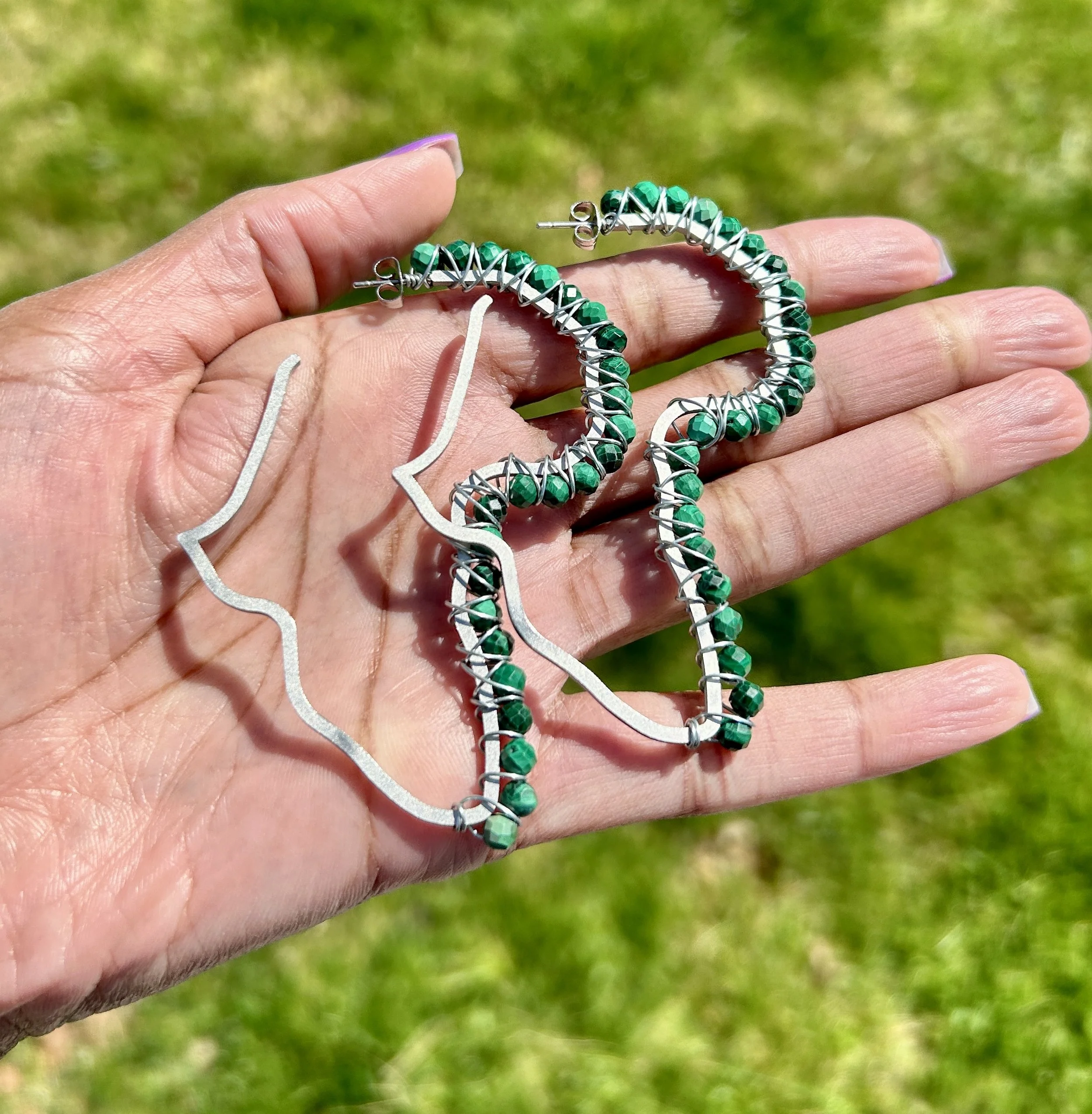 Hand holding a pair of earrings with silver and green beads, outdoors on grass.