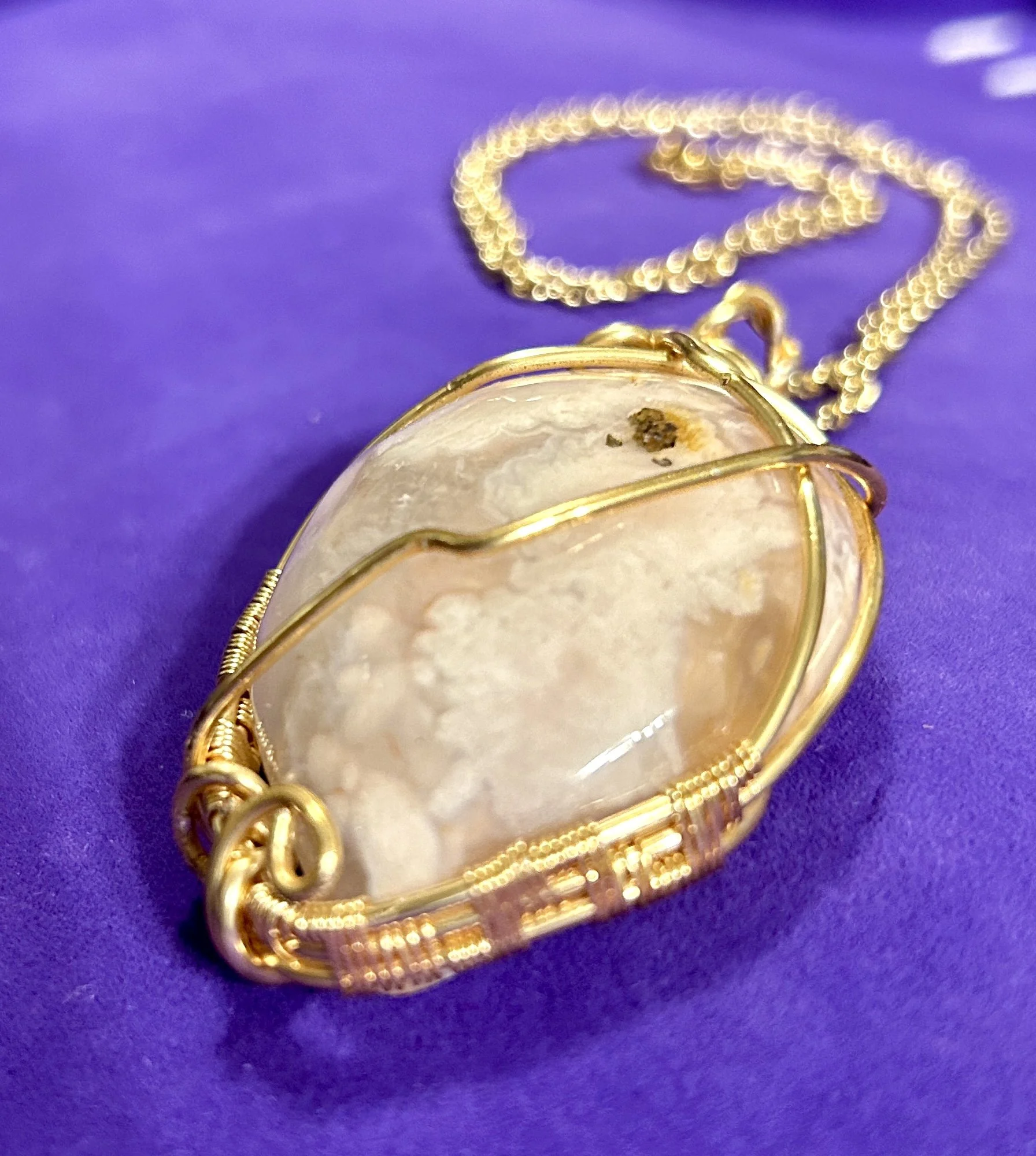 A pendant necklace with a large oval-shaped white stone wrapped in gold wire, resting on a purple surface.