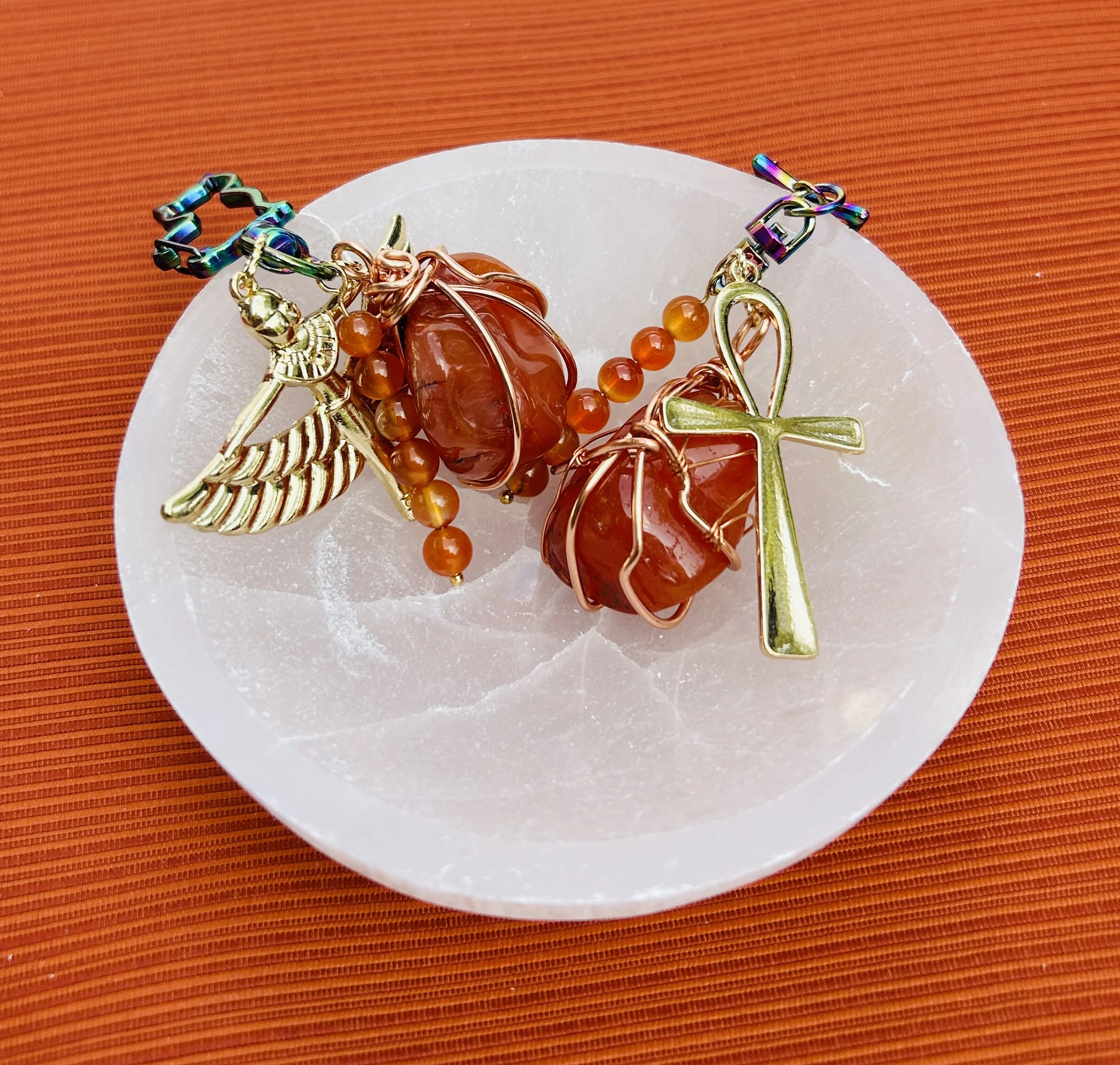 A bracelet with charms, including an angel wing, a cross, and two large orange stones, placed on a white circular surface on an orange background.