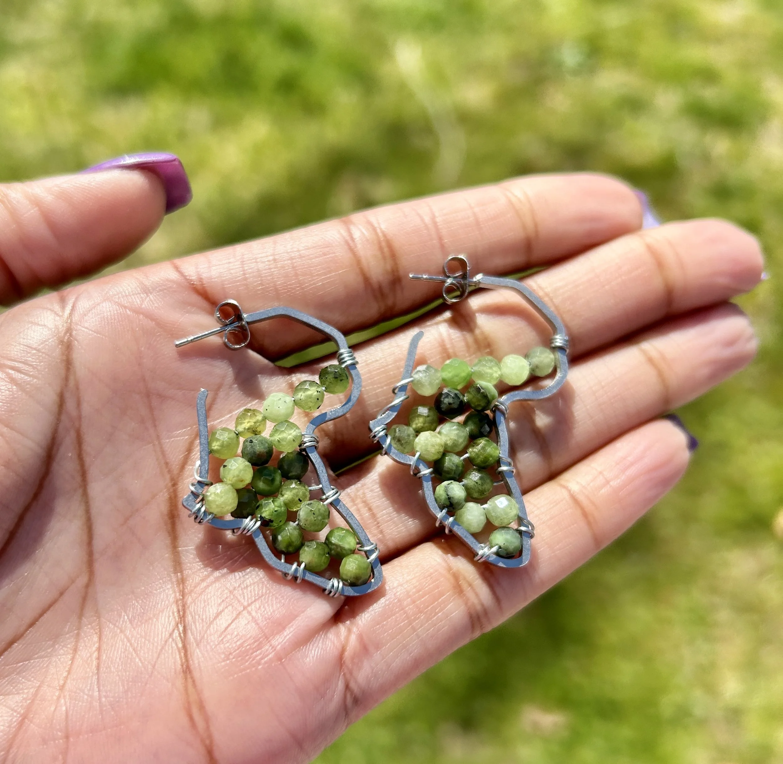Hand holding a pair of earrings made with green and black beads and metal wire, outdoors on grass.