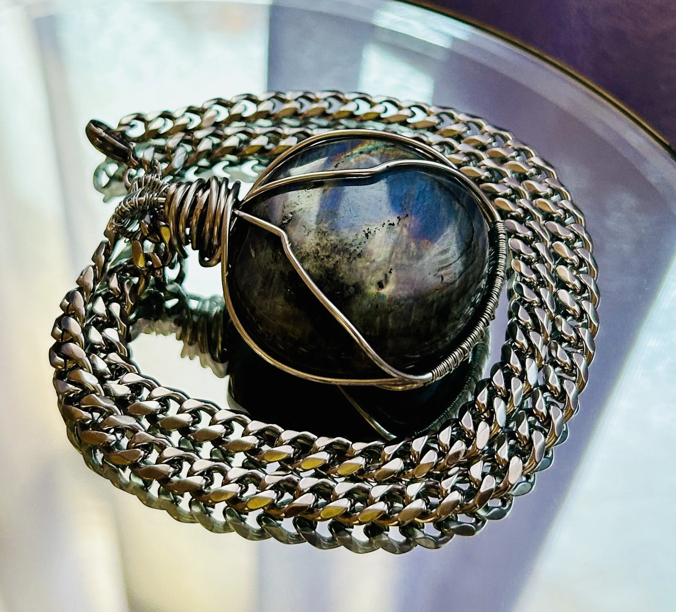 Close-up of a black stone pendant wrapped in wire, resting on a silver chain, placed on a reflective surface with a blurred background.