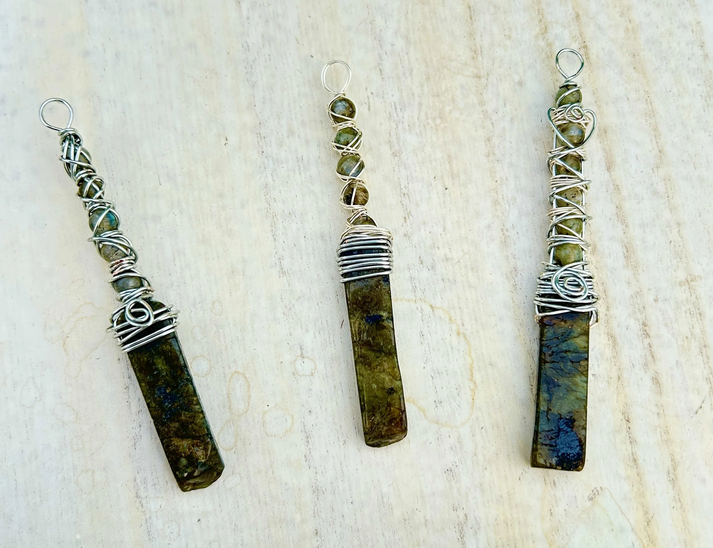Three handmade wire-wrapped necklaces with long, rectangular, multicolored stone pendants on a light-colored surface.