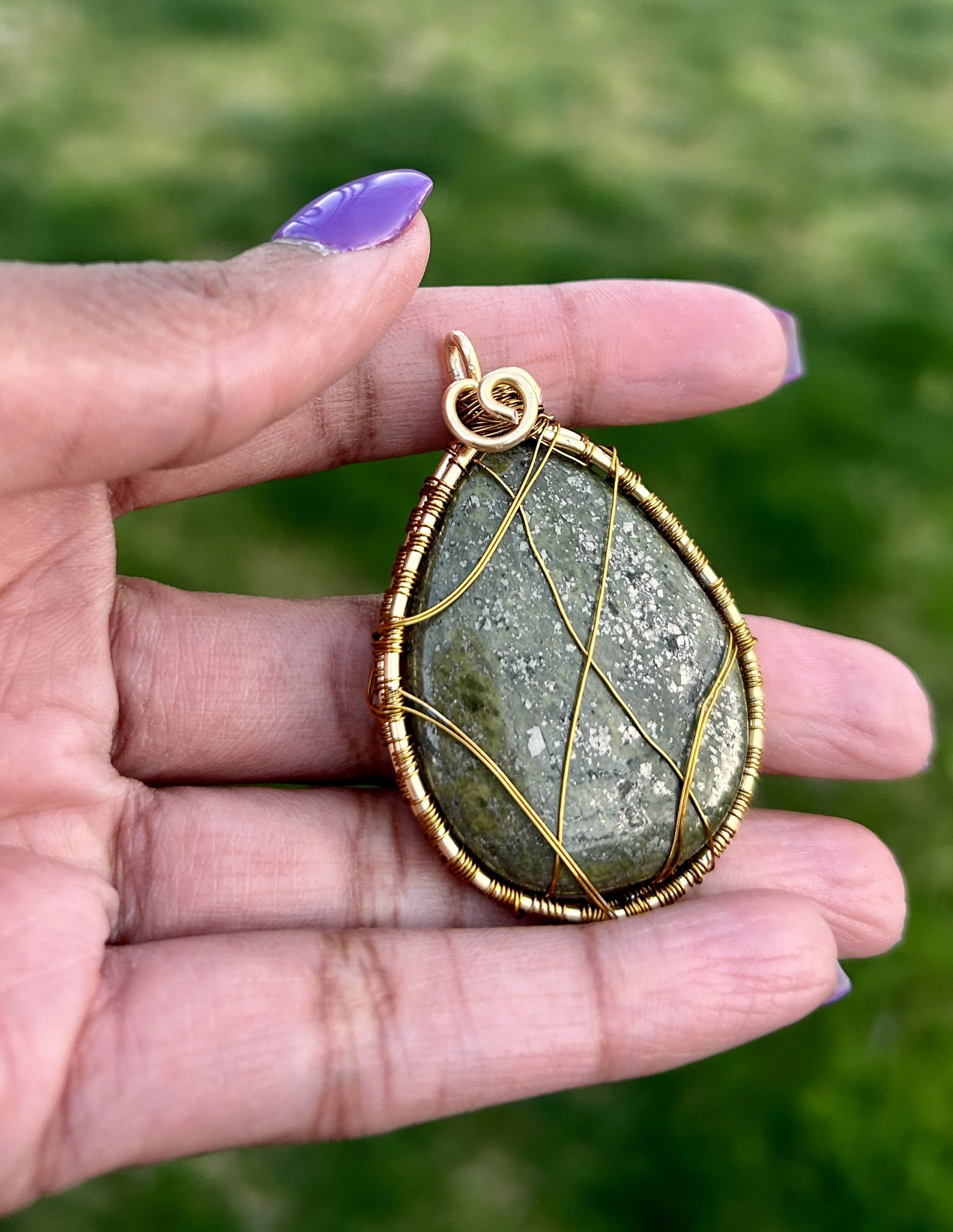 A hand with lavender-colored nails holding a pendant with a greenish stone wrapped in gold wire, set against a green background.