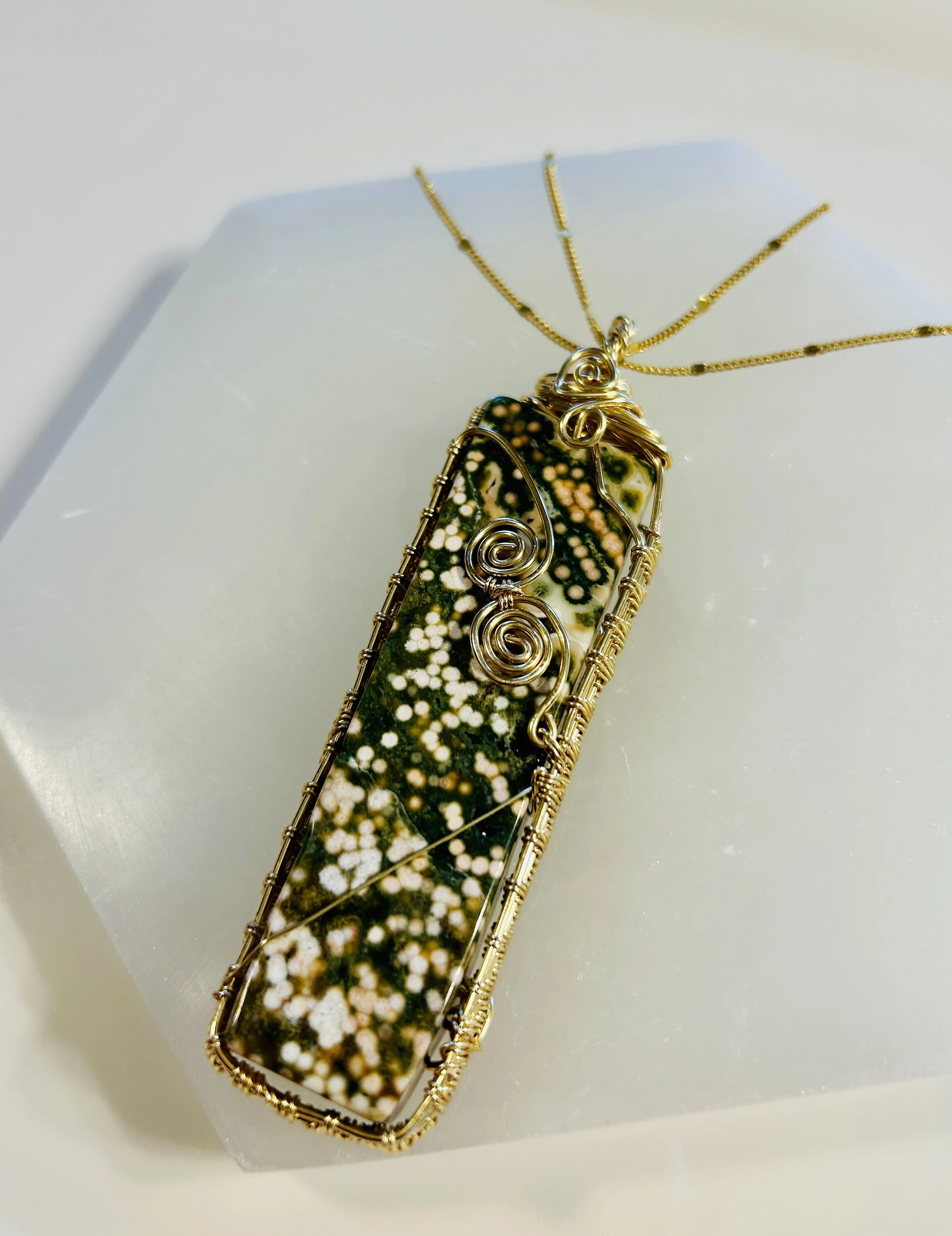 A pendant necklace featuring a large, rectangular, glass-encased specimen with black, green, and white patterns, wrapped in gold-colored wire with decorative wire spirals, hanging from a gold chain.