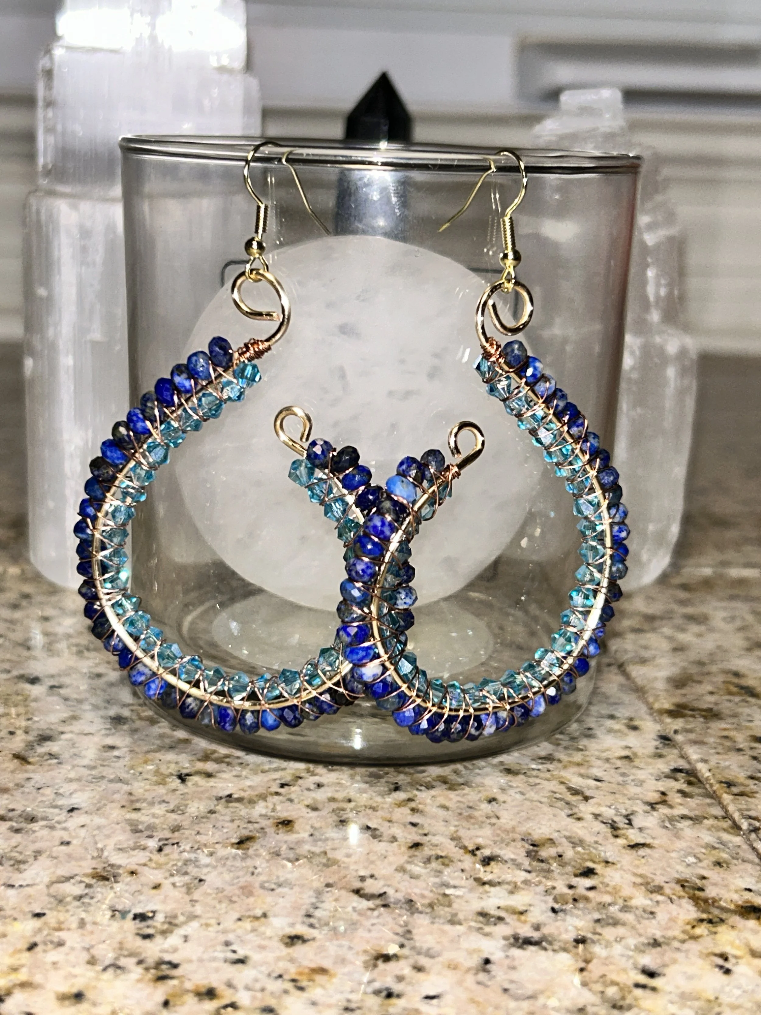 Pair of blue and teal beaded hoop earrings hanging over a glass candle holder on a speckled countertop.