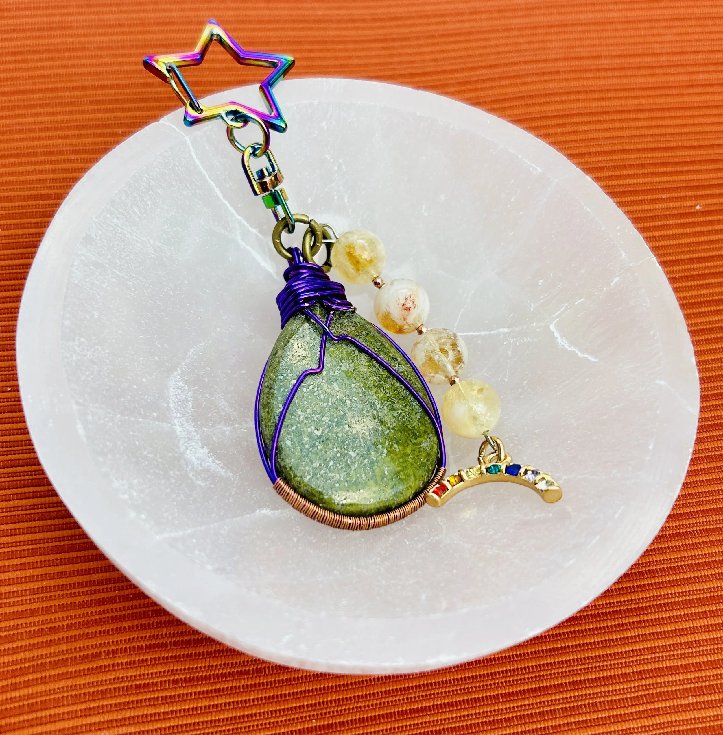 A handcrafted necklace with a large green teardrop-shaped stone wrapped in purple wire, featuring a star-shaped clasp, white beads, and a small rainbow crescent pendant, displayed on a white circular plate with an orange background.