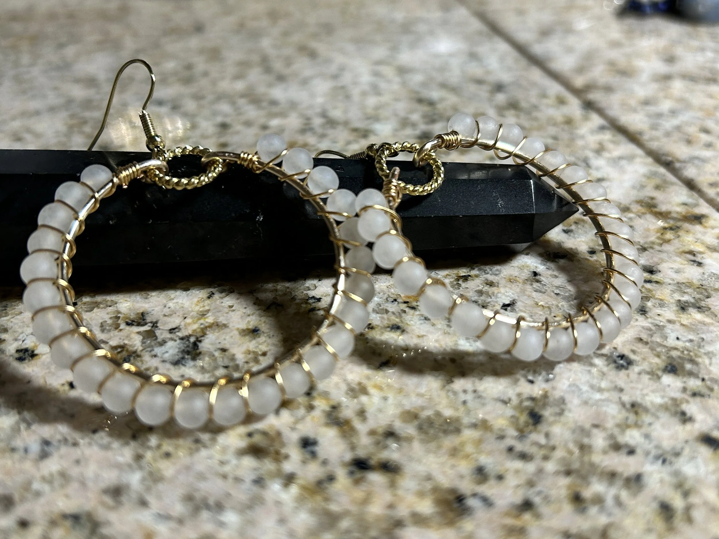 A pair of hoop earrings with white beads and gold wire on a black obelisk-shaped object on a speckled countertop.