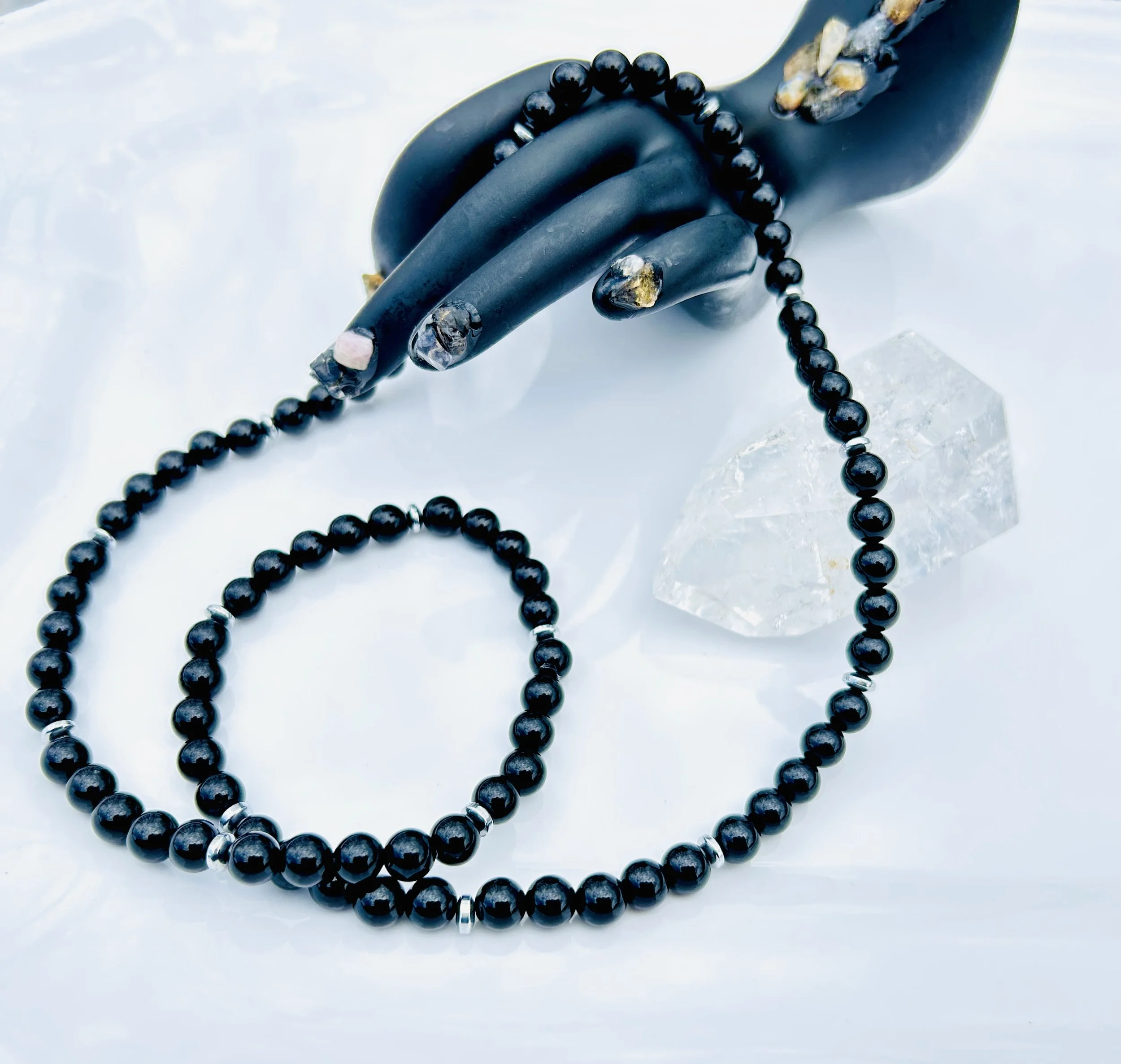 A black beaded necklace with silver accents, a black hand-shaped jewelry holder with gold details, a clear quartz crystal, and a pink stone on a white surface.