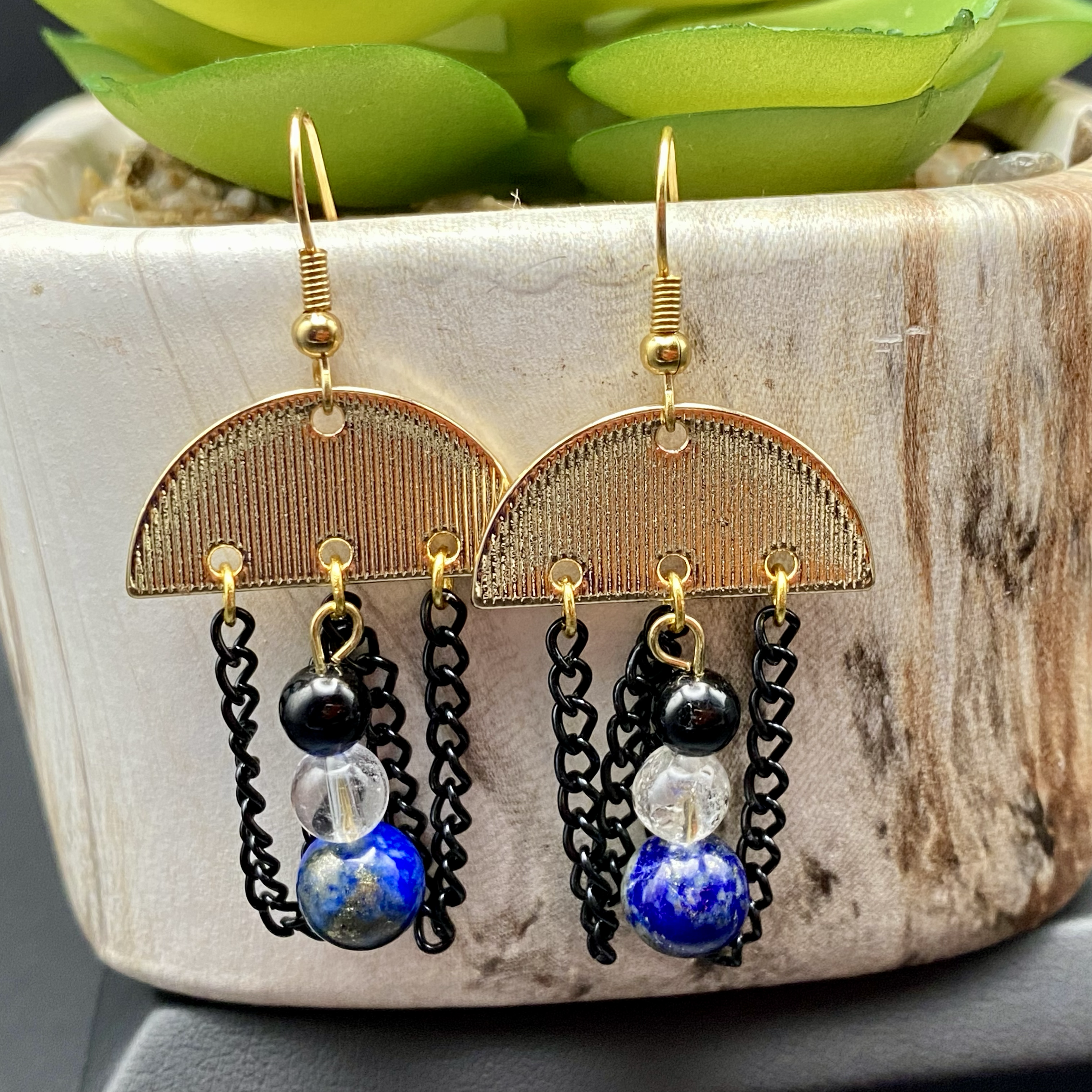 Gold and black dangle earrings with chain detailing and spherical beads, displayed on a textured beige surface in front of a green succulent plant.