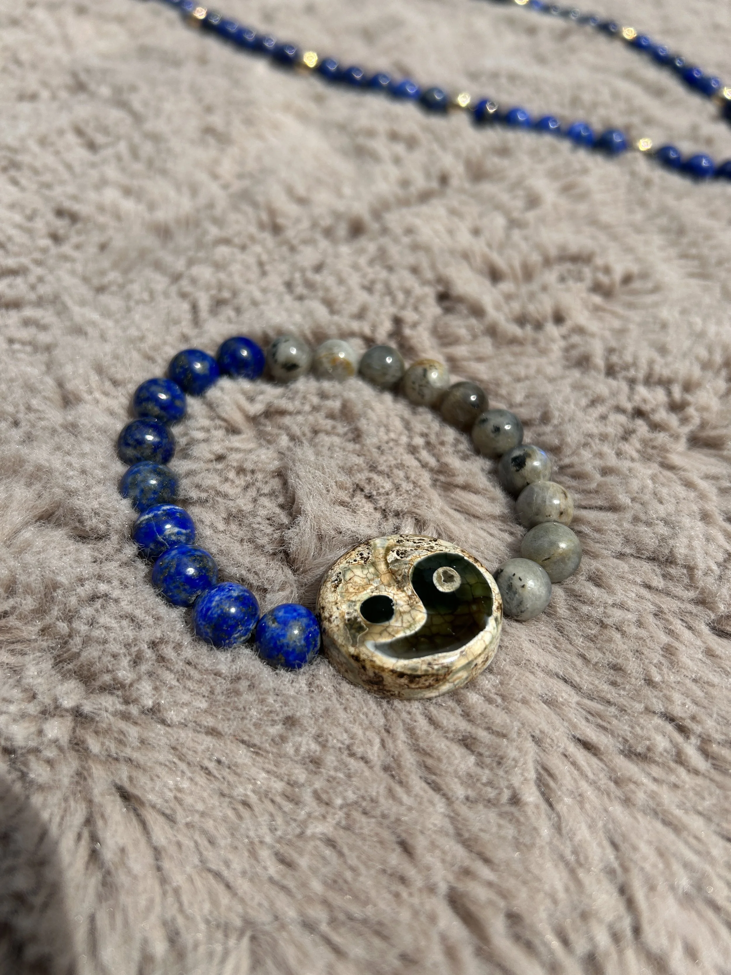 A beaded necklace with alternating blue and gray beads, featuring a yin-yang pendant with black and white stone elements, resting on a soft, beige furry surface.