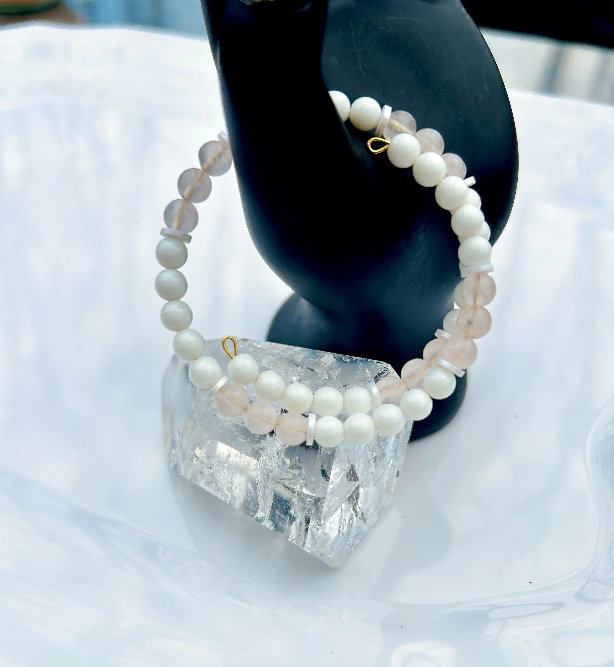 A pearl and light pink beaded bracelet draped over a black mannequin hand, resting on a clear glass block on a white surface.