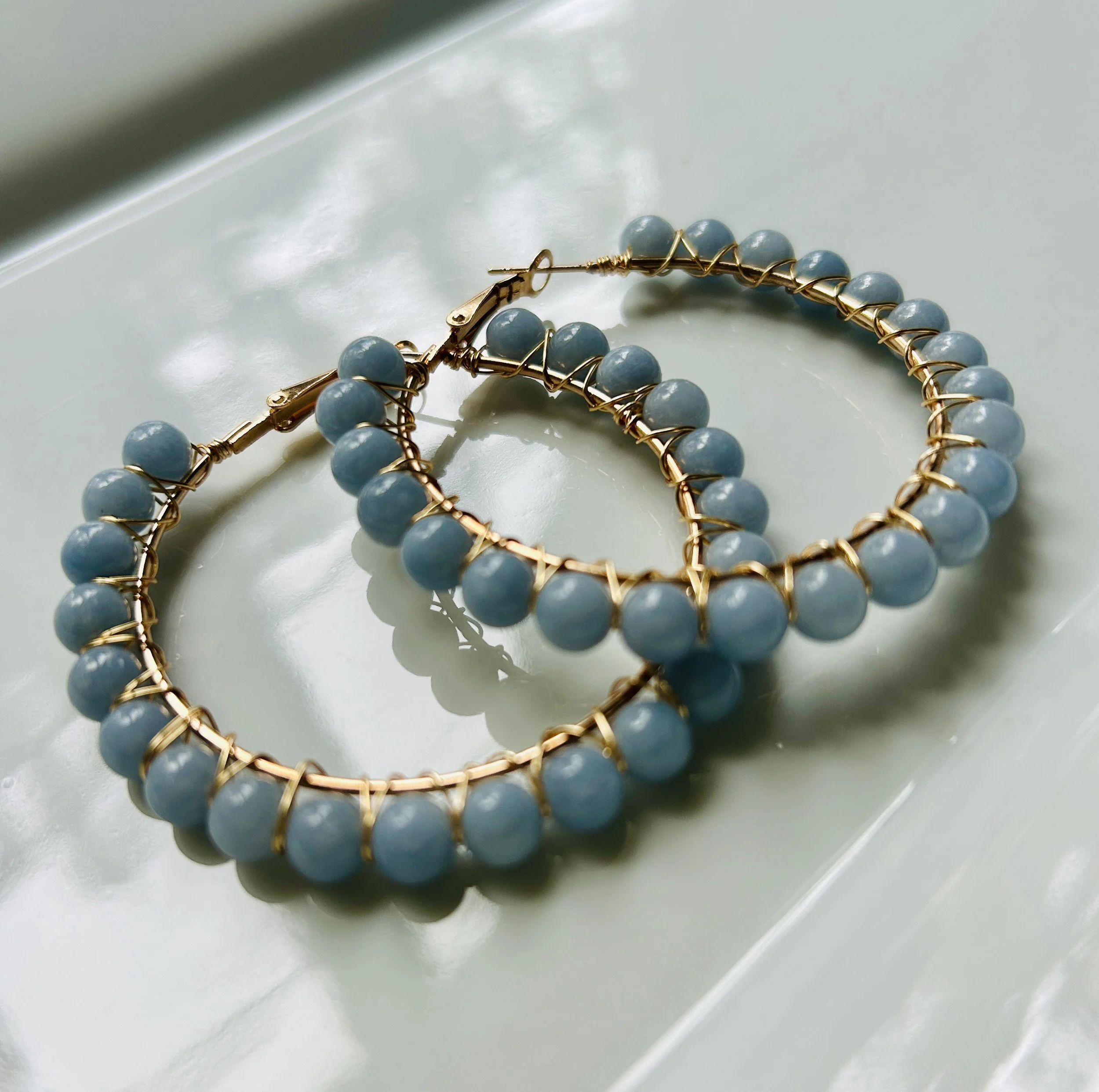 A pair of gold hoop earrings with light blue beads placed on a reflective surface.