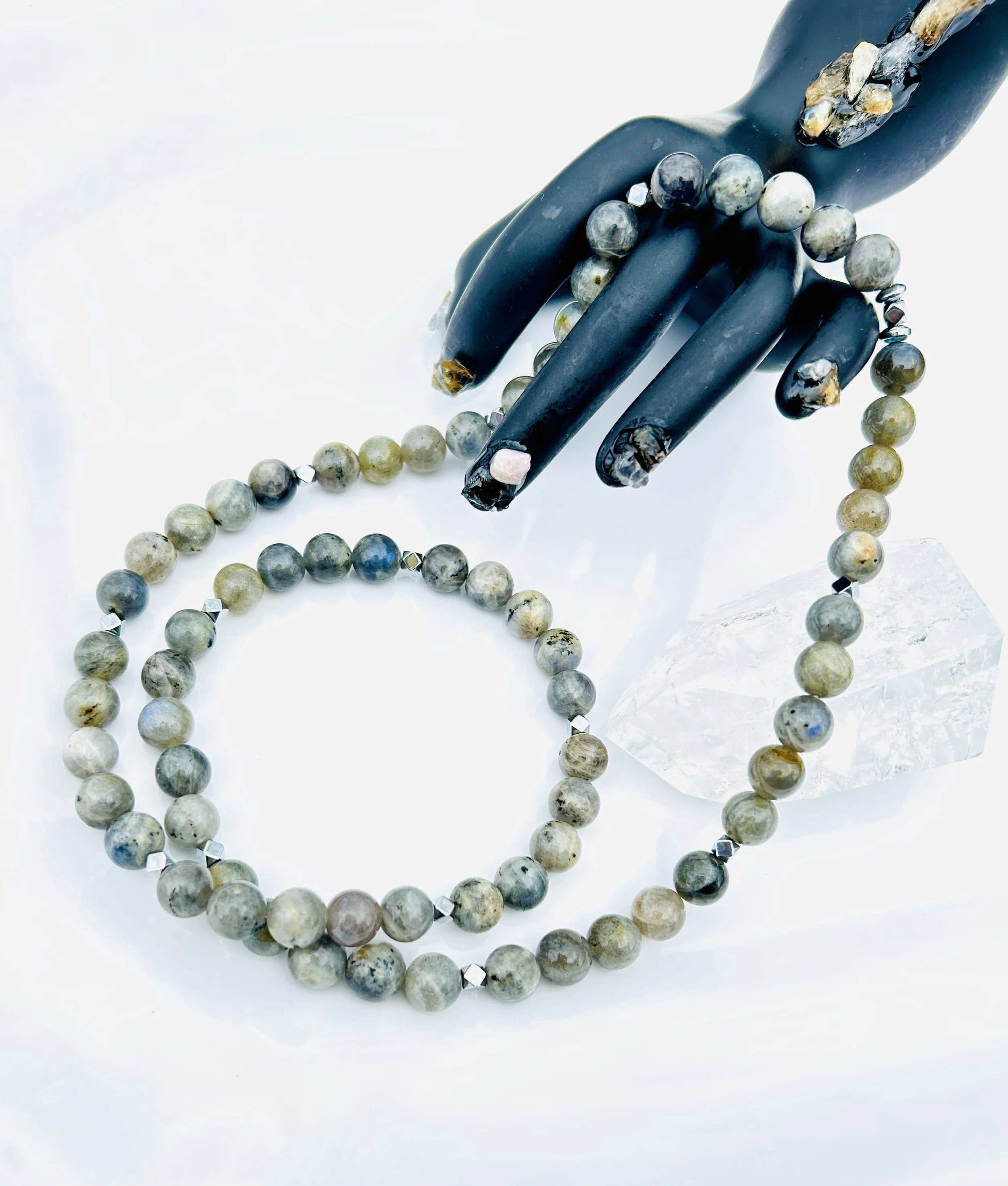 A black hand sculpture holding a beaded necklace with round and faceted stones, next to a clear quartz crystal on a white surface.
