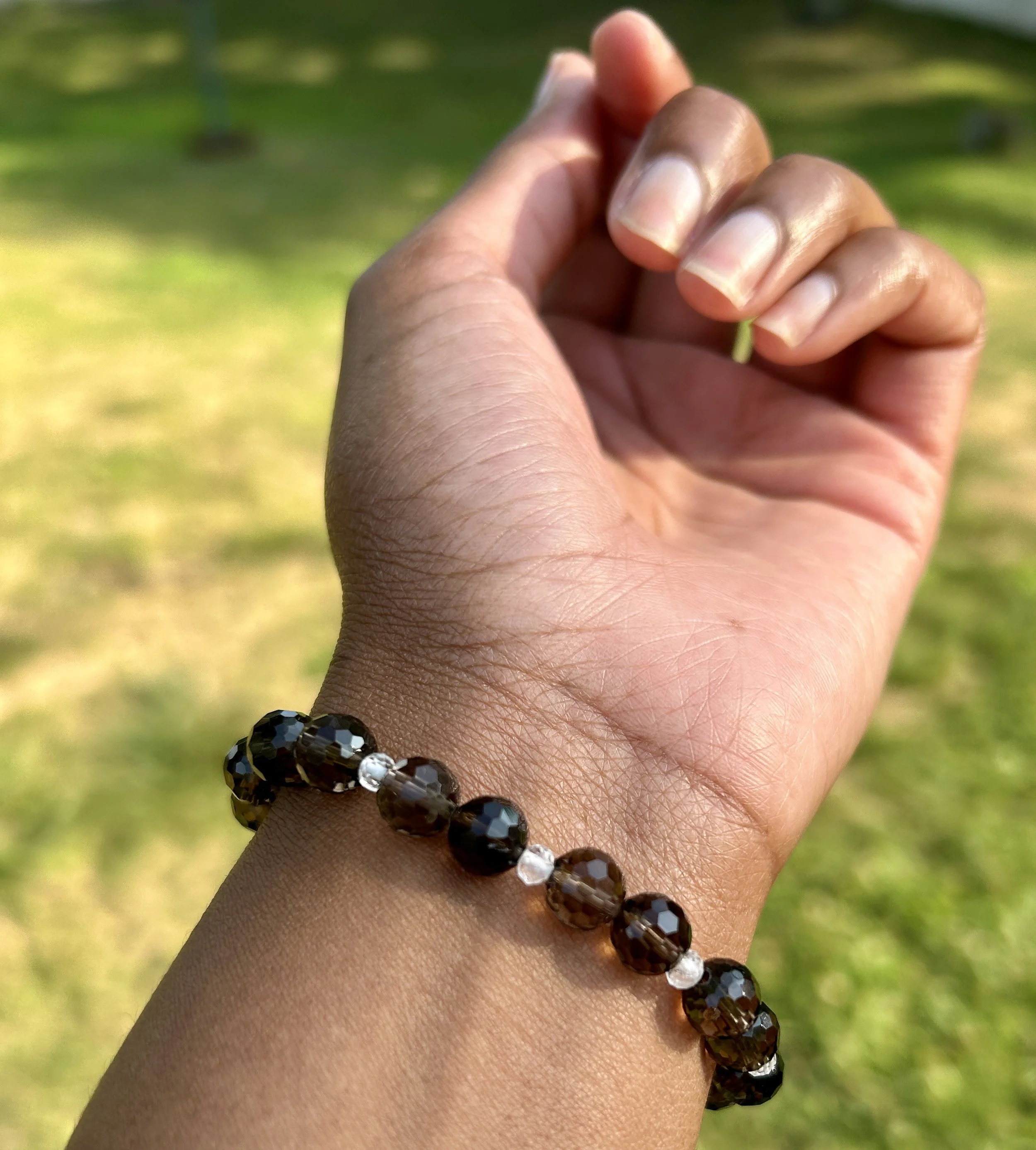 A person's hand wearing a black beaded bracelet with silver accents, shown outdoors with a grassy background.