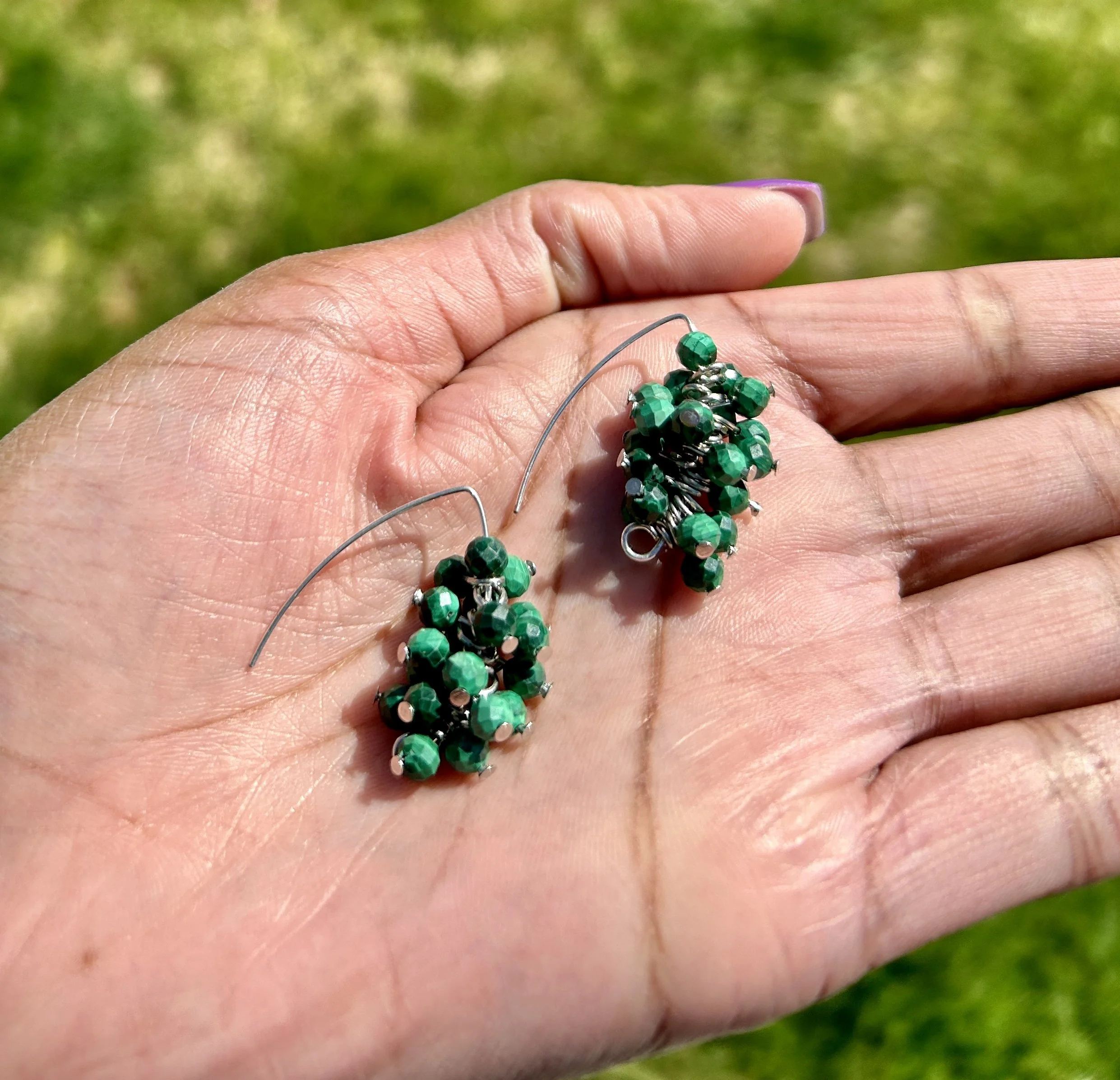 Green beaded dangling earrings held in a person's hand outdoors.