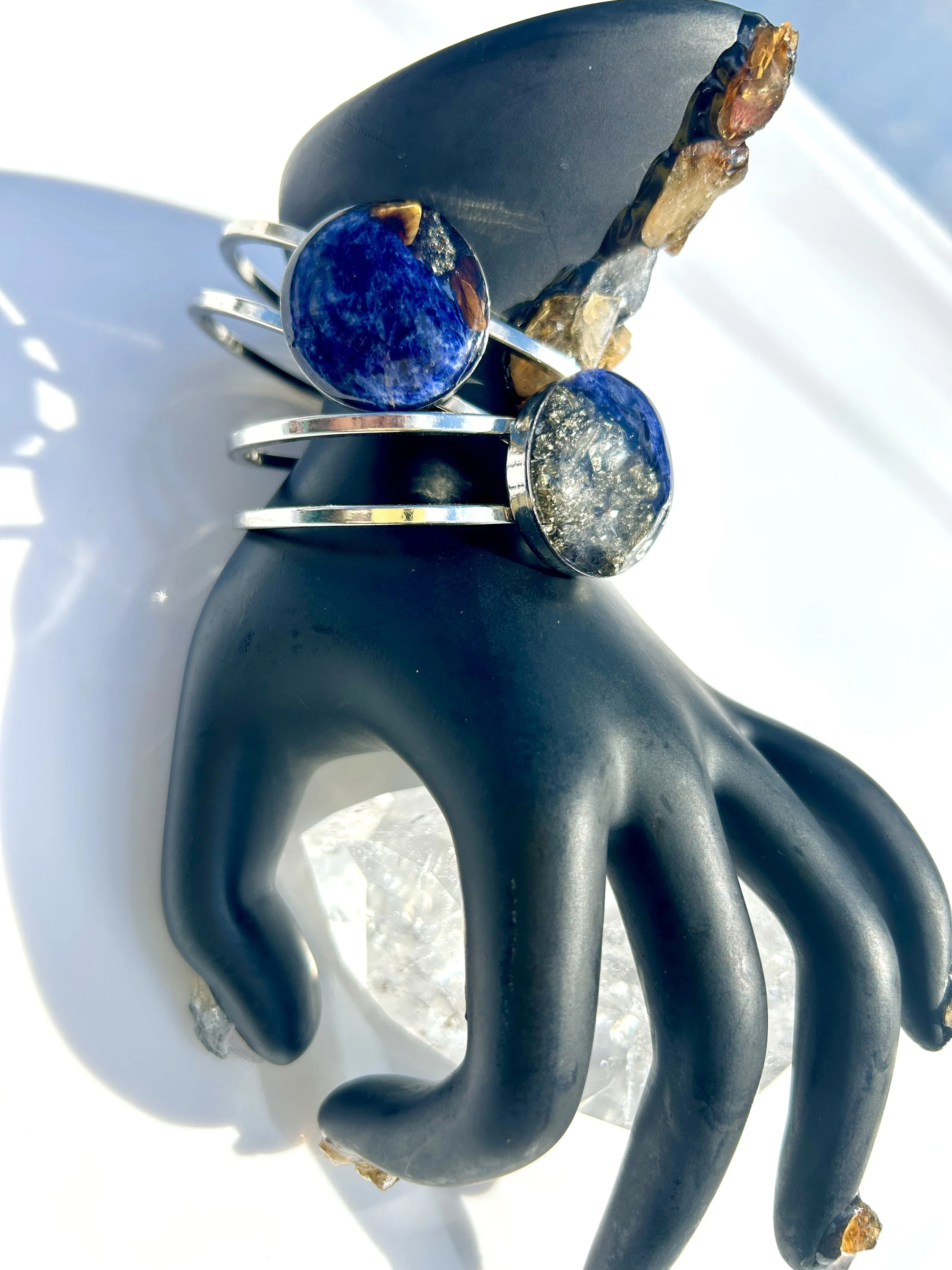 Two silver rings with large blue and silver textured stones resting on a black hand-shaped jewelry display stand. The display stand is placed on a clear crystal base, with sunlight casting shadows.