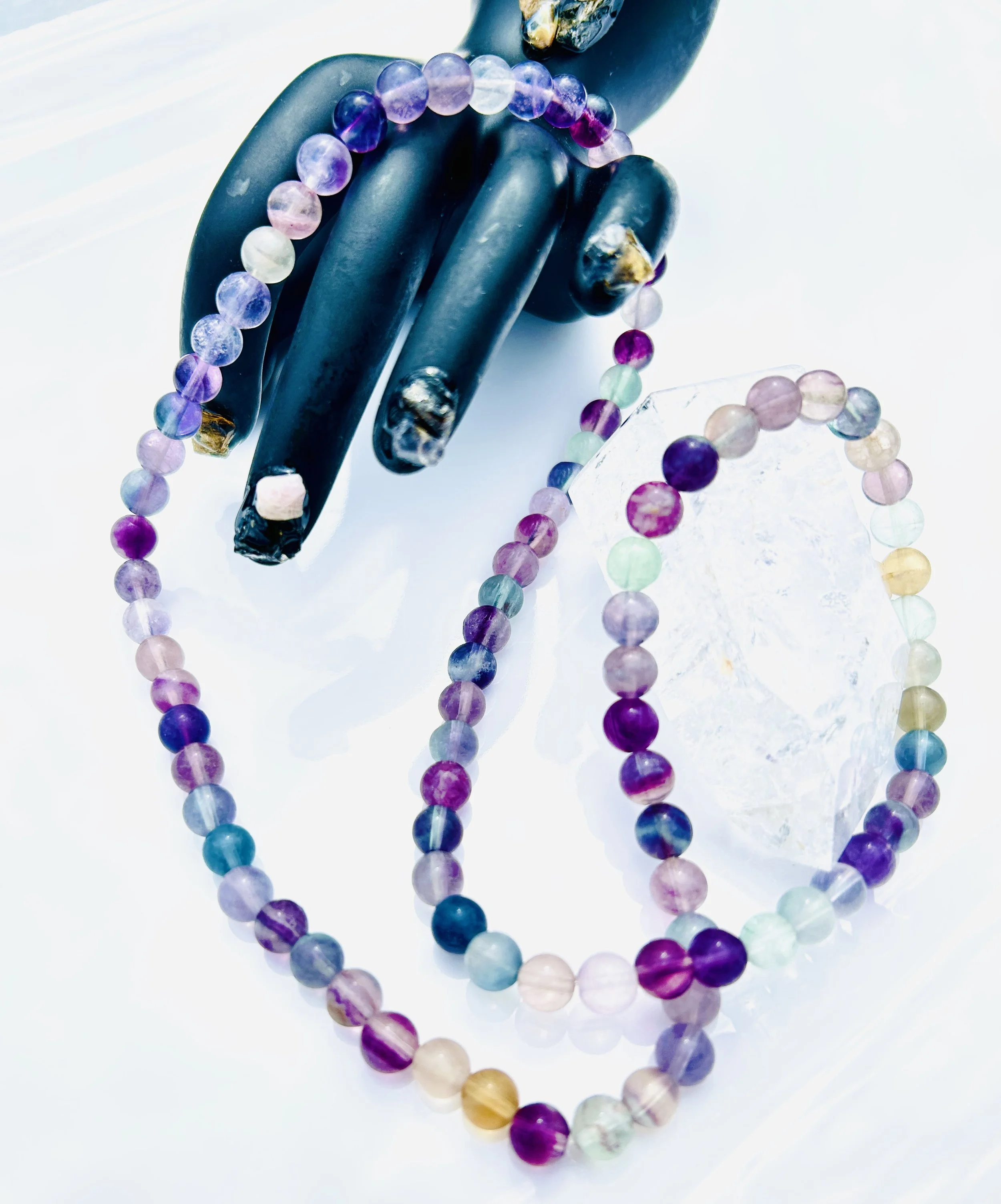 Colorful beaded necklaces with a purple, pink, green, and blue color scheme, draped over black gloves and a clear crystal, against a white background.