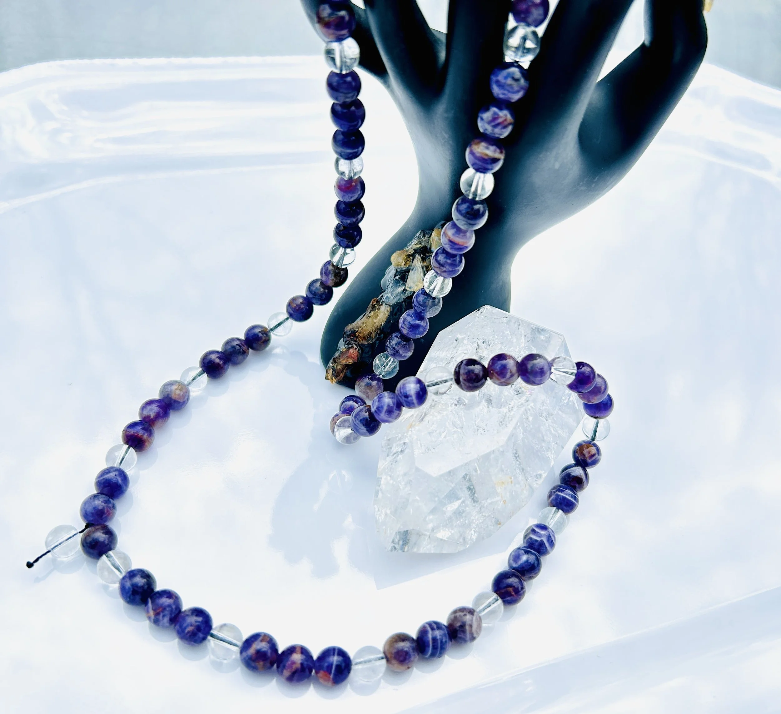 A black mannequin hand displays a purple and clear beaded necklace draped over fingers, with a loose strand on a white surface, and a large clear crystal behind the hand.