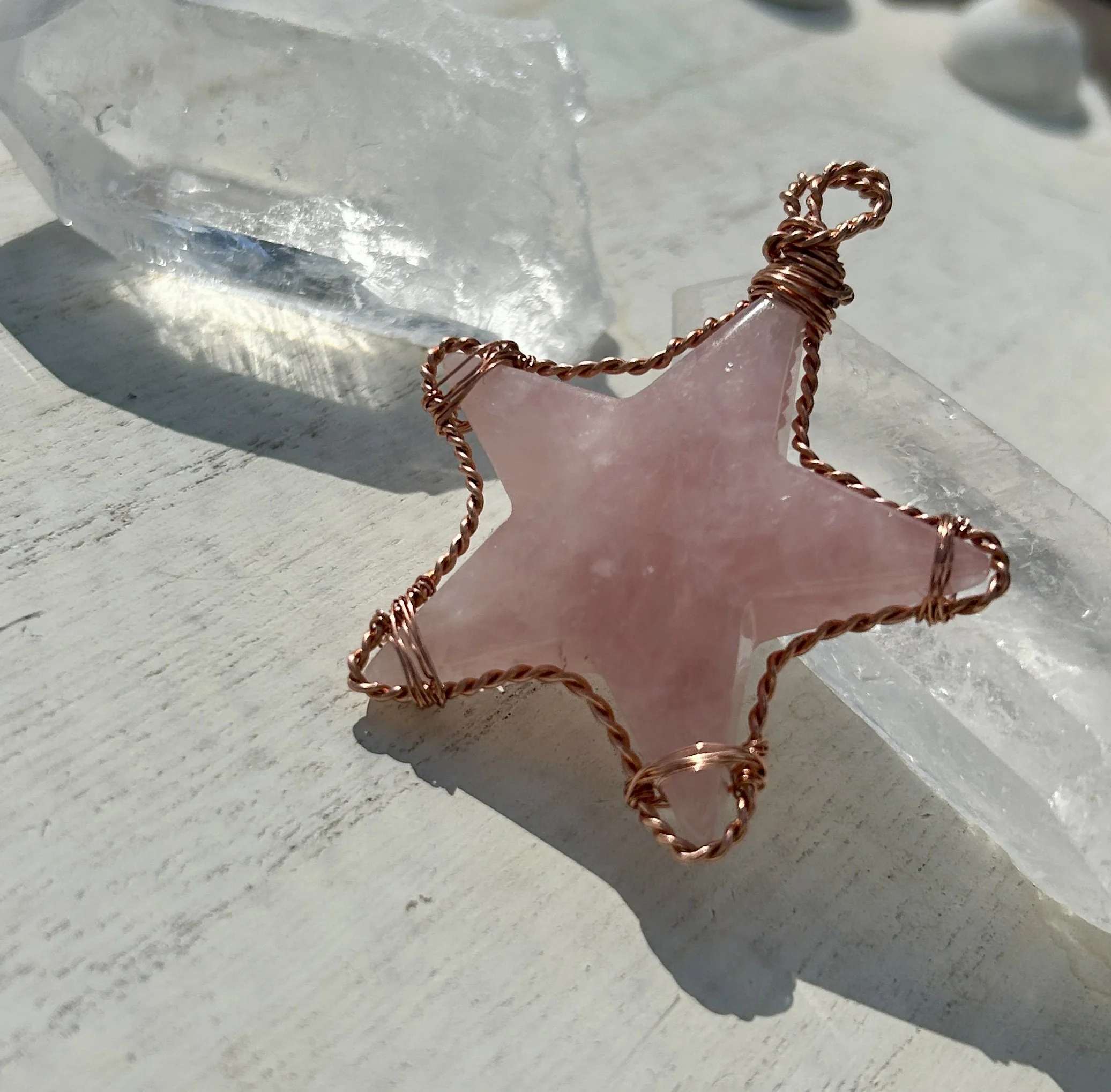A pink star-shaped pendant wrapped with rose gold wire, resting on a clear, chunky crystal on a white wooden surface.